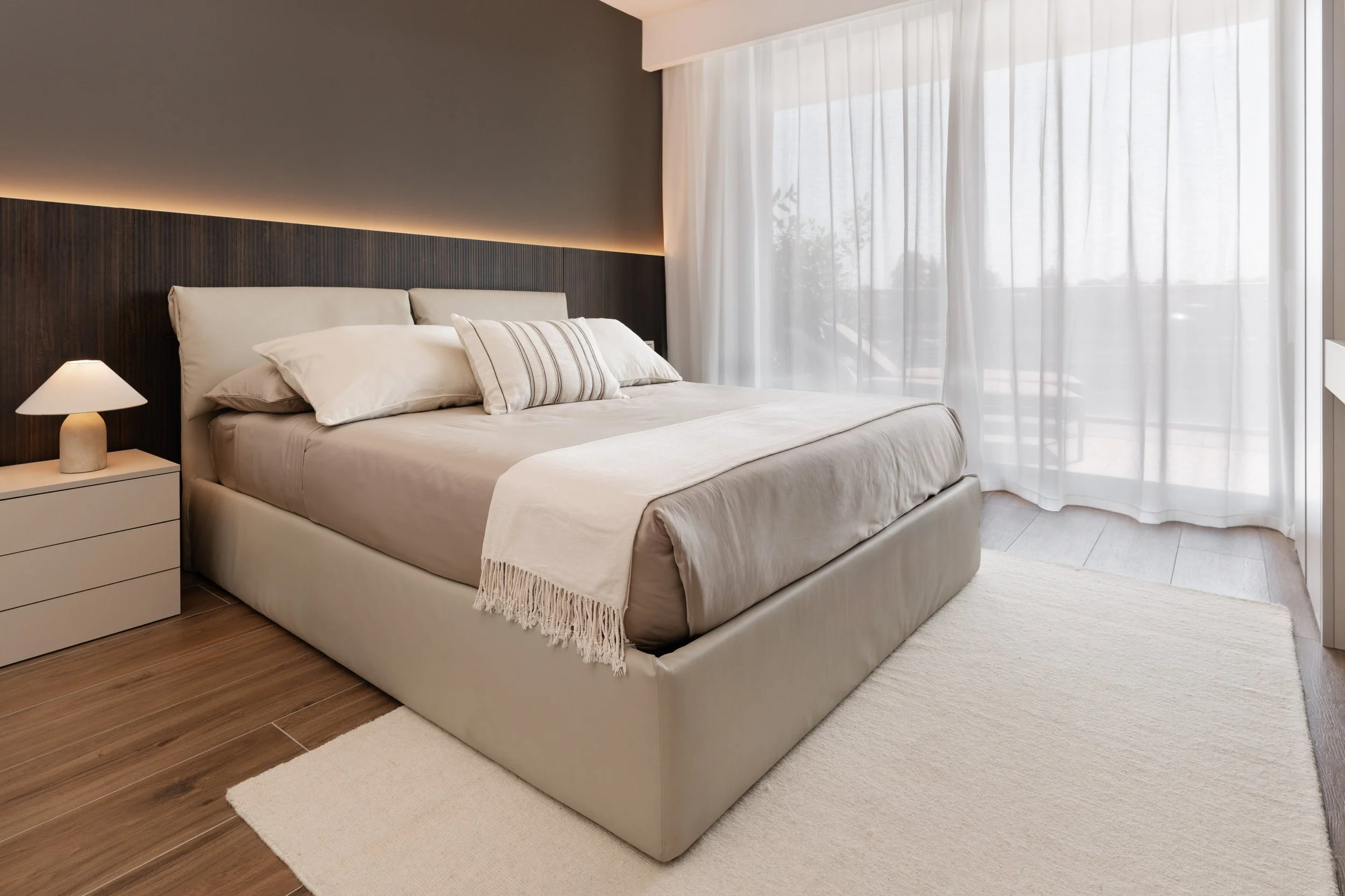 Stanza da letto elegante con letto matrimoniale, cuscini, copriletto beige, tappeto bianco, lampada da tavolo e tende bianche trasparenti, con luce naturale che entra dalla finestra.