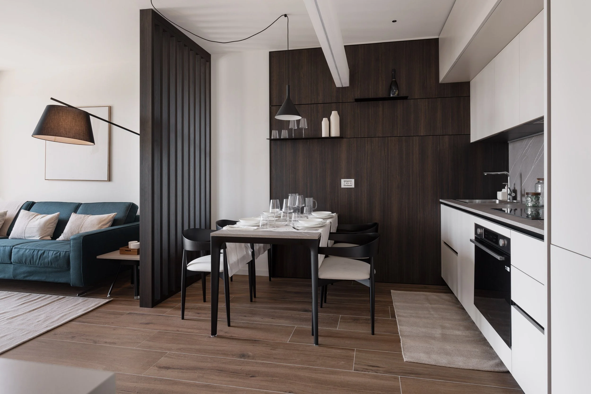 Interior di un soggiorno e angolo cottura con tavolo apparecchiato per pasti, divano blu con cuscini, lampada nera a soffitto e pareti di legno scuro e bianco.