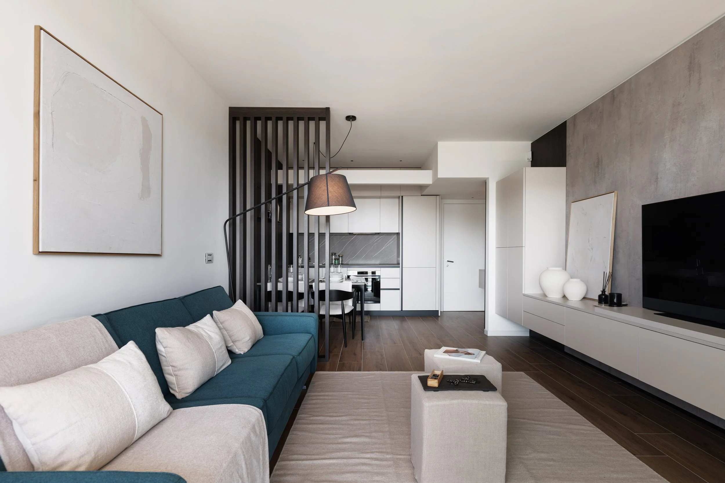 Interno di un soggiorno moderno con divano blu, pareti bianche e grigie, tavolino bianco, TV e decorazioni minimaliste, con cucina aperta sullo sfondo.