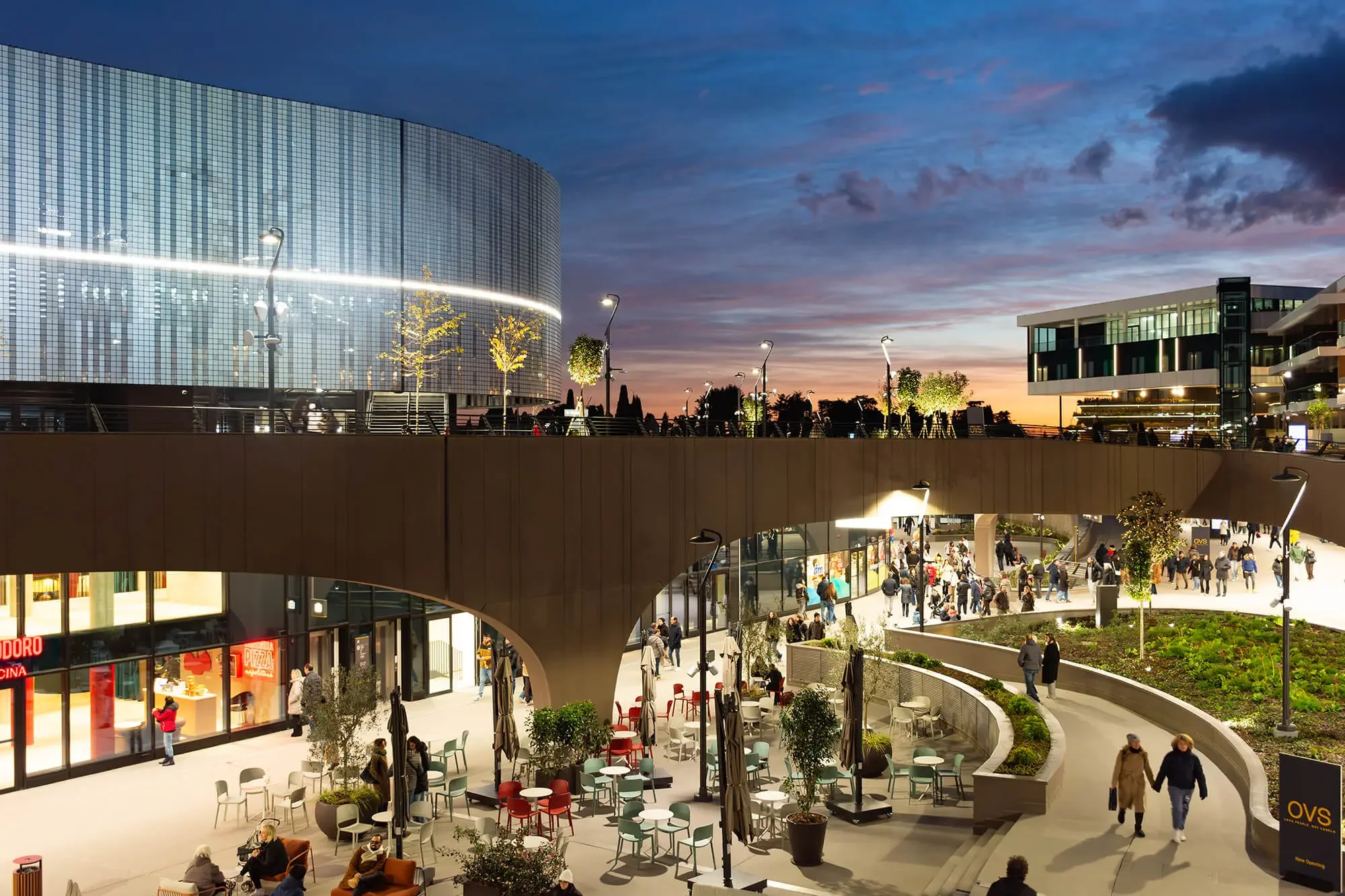 Scalo commerciale all'aperto al tramonto con persone che camminano, sedie e tavoli, edifici moderni illuminati, alberi e luci stradali.