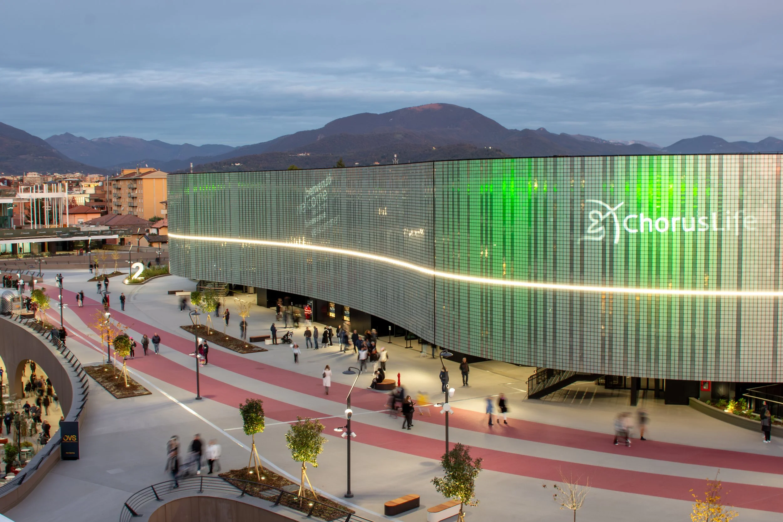Immagine di un centro commerciale moderno con una grande facciata a schermo di LED che mostra il logo di Chorus e altri segnali digitali, con molte persone che passeggiano nel piazzale e alberi piantati lungo il percorso.