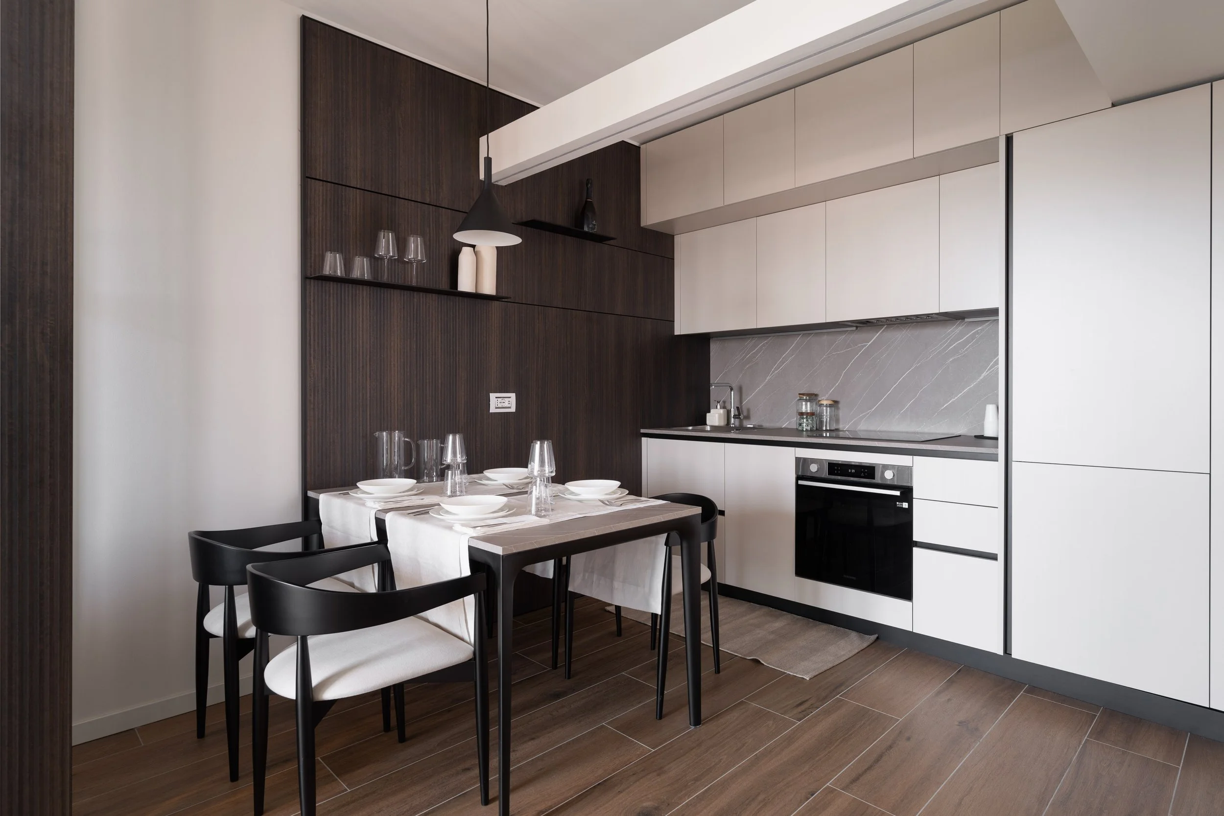 Cucina moderna con mobili bianchi, superfice in pietra grigia, tavolo apparecchiato con tovaglia bianca, quattro bicchieri e piatti, sedie nere con seduta bianca, pareti in legno scuro, illuminazione con lampada nera a sospensione.