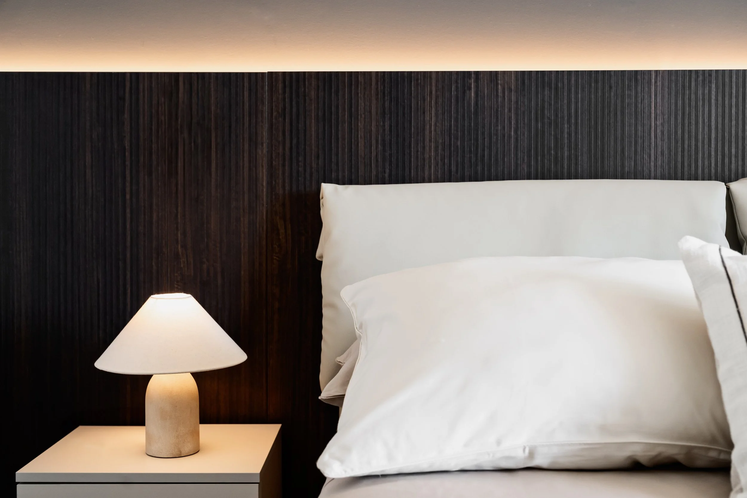 Una camera da letto elegante con un letto con copripiumino bianco e cuscini abbinati, una parete in legno scuro e una lampada da tavolo bianca sul comodino vicino al letto.