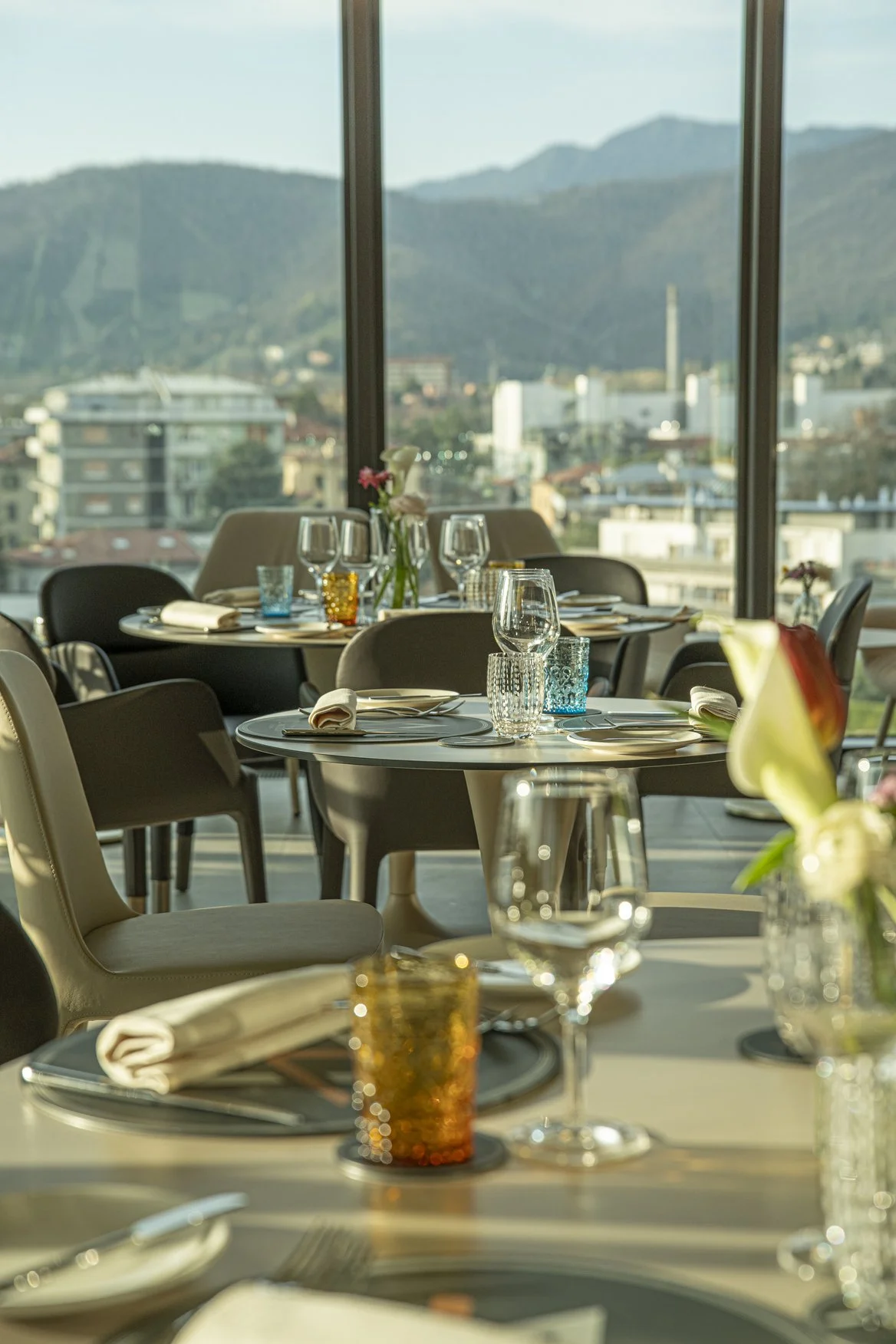 Tavoli pronti con bicchieri, piatti, posate e tovaglioli in un ristorante con grandi finestre e vista sulle montagne.