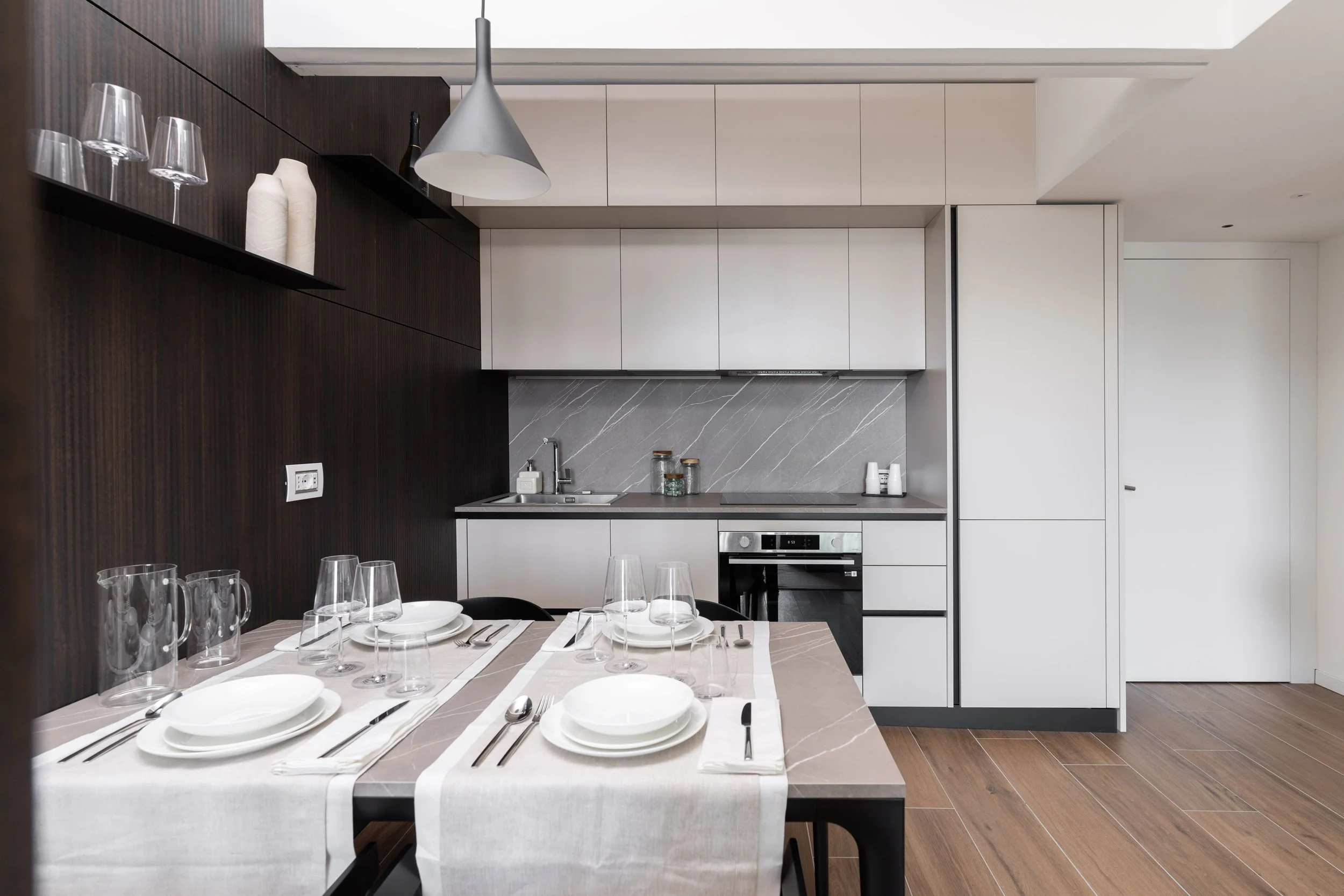 Cucina moderna con tavolo apparecchiato per quattro persone, armadietti bianchi, parete in marmo chiaro, pavimento in legno, illuminazione a soffitto.