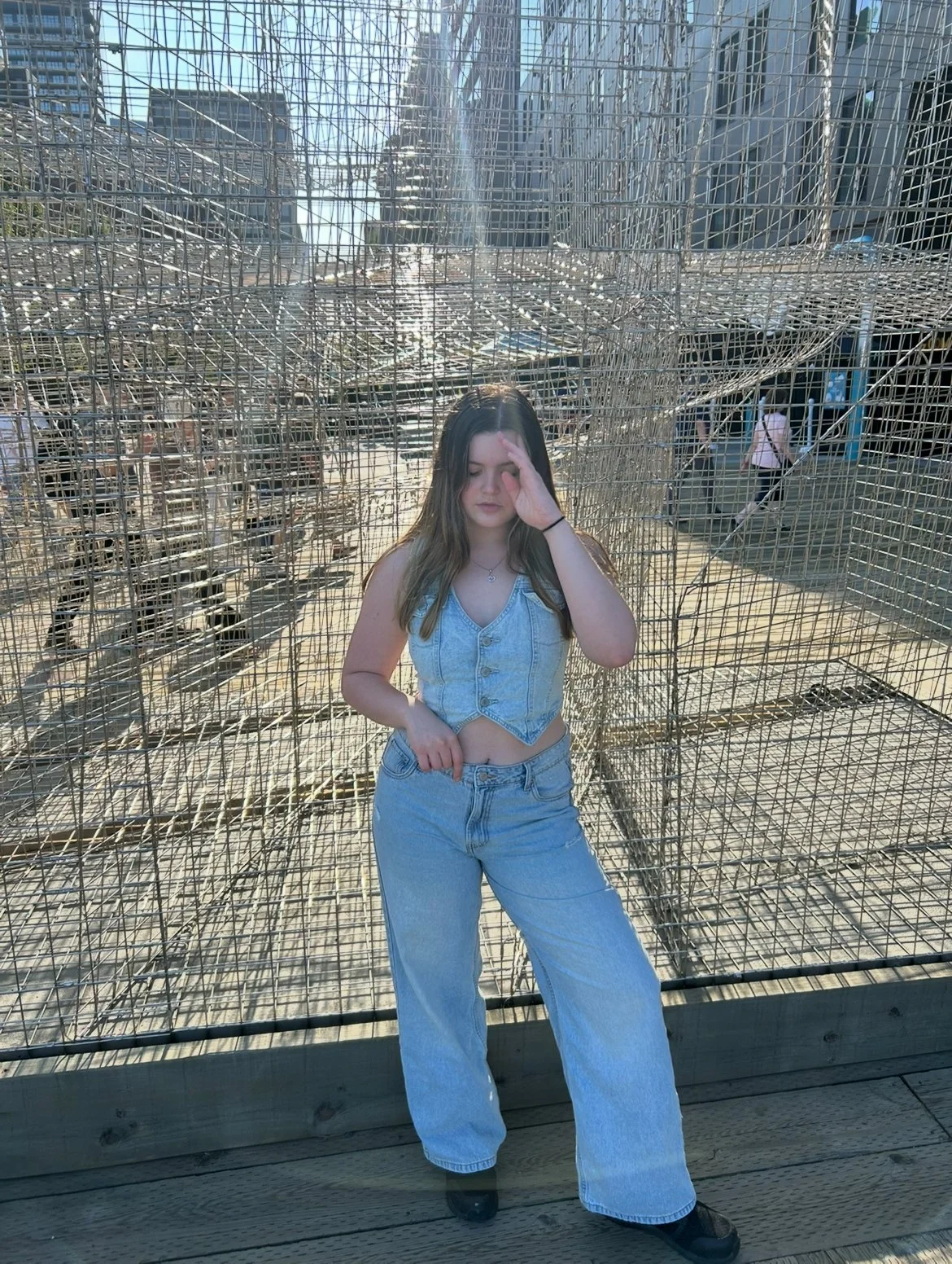 A young woman with long brown hair in a sleeveless denim vest and jeans standing inside a wire cage structure, with her eyes closed and one hand touching her face, outdoors with urban buildings in the background.
