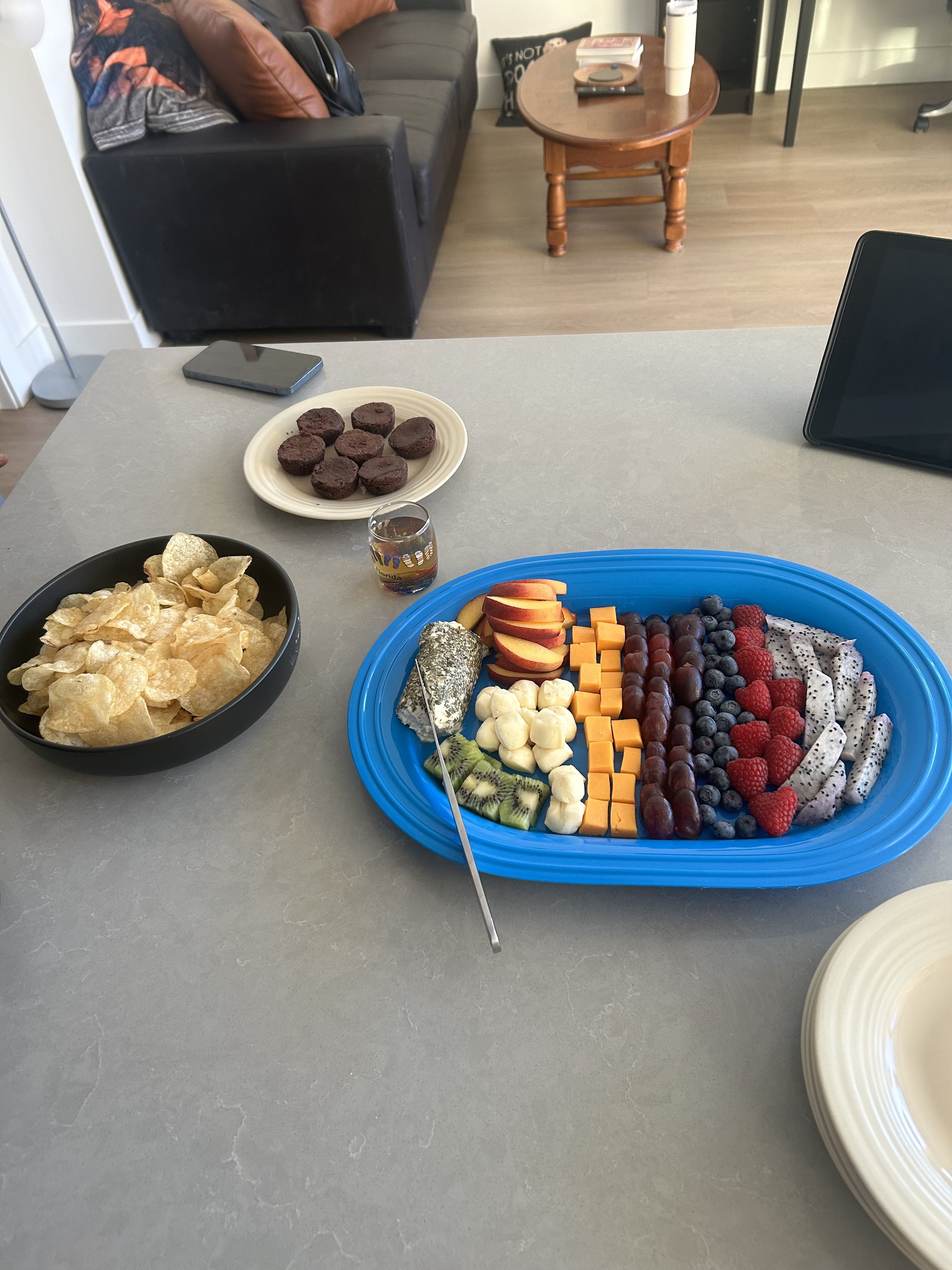 A table with a large blue platter of assorted sliced and whole fruit, a black bowl of potato chips, a white plate with dark cookies, and a small glass of dark liquid. In the background, there is a black phone on the table and a living room with a dar