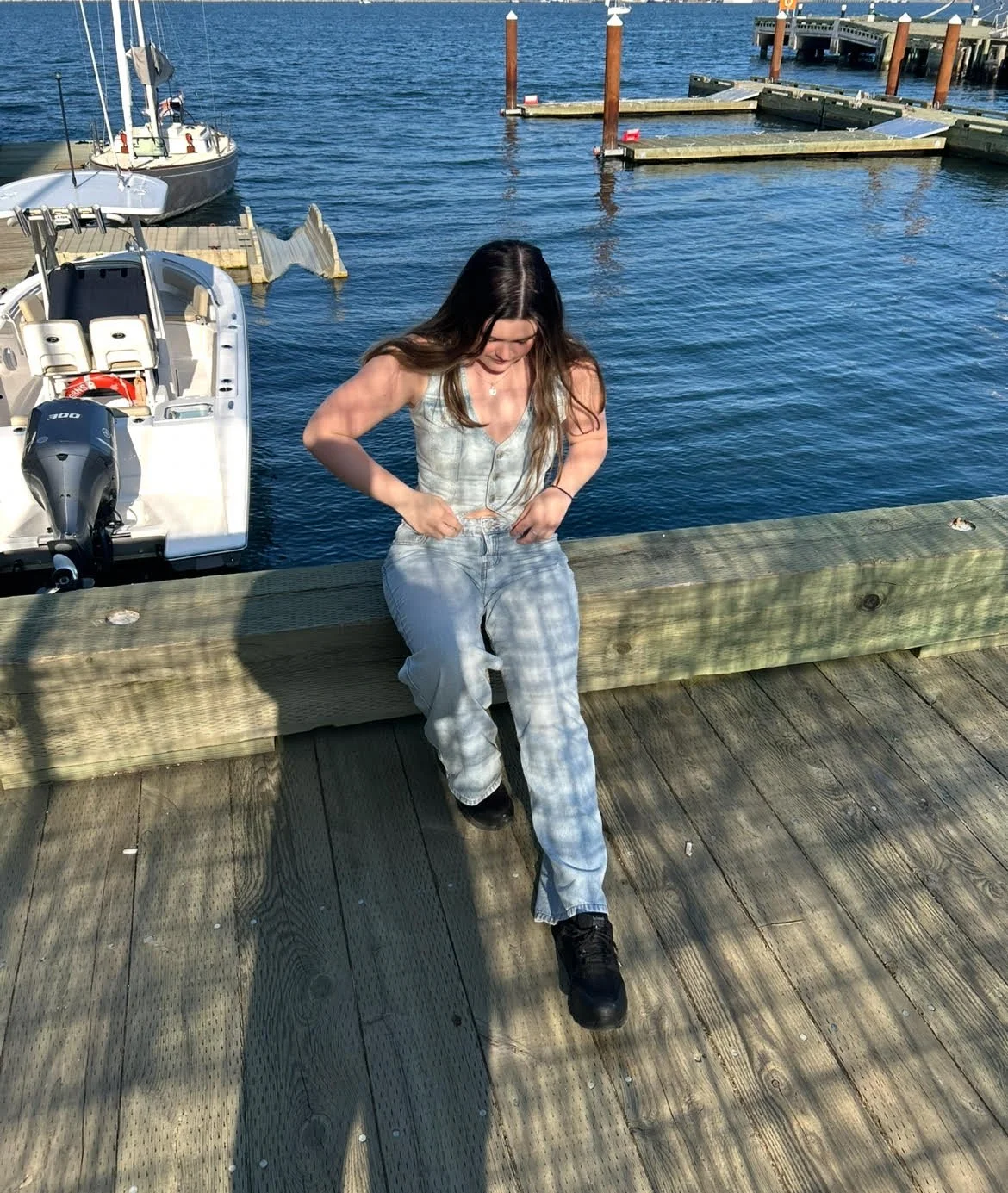 Abi Mack with a full denim outfit sitting on a dock with a boat and water behind her, the water is a beautiful blue.