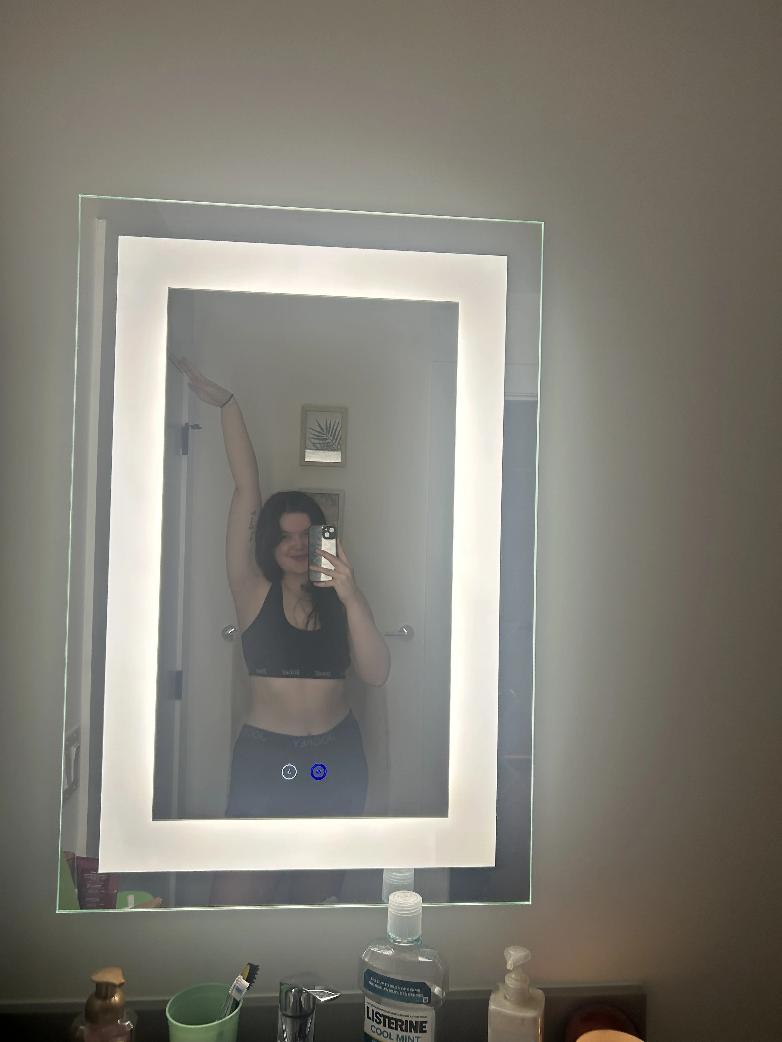 Abi Mack taking a mirror selfie in a bathroom. She is wearing a black sports bra and black shorts, with one arm raised above her head. The bathroom has a backlit mirror and various toiletries on the counter.