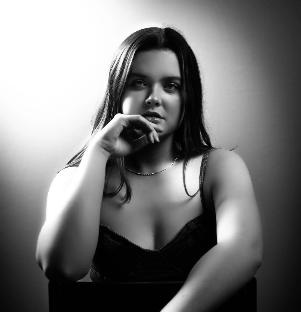 Abi Mack sitting on a stool looking at a camera with her hand near her chin, image is in black and white.