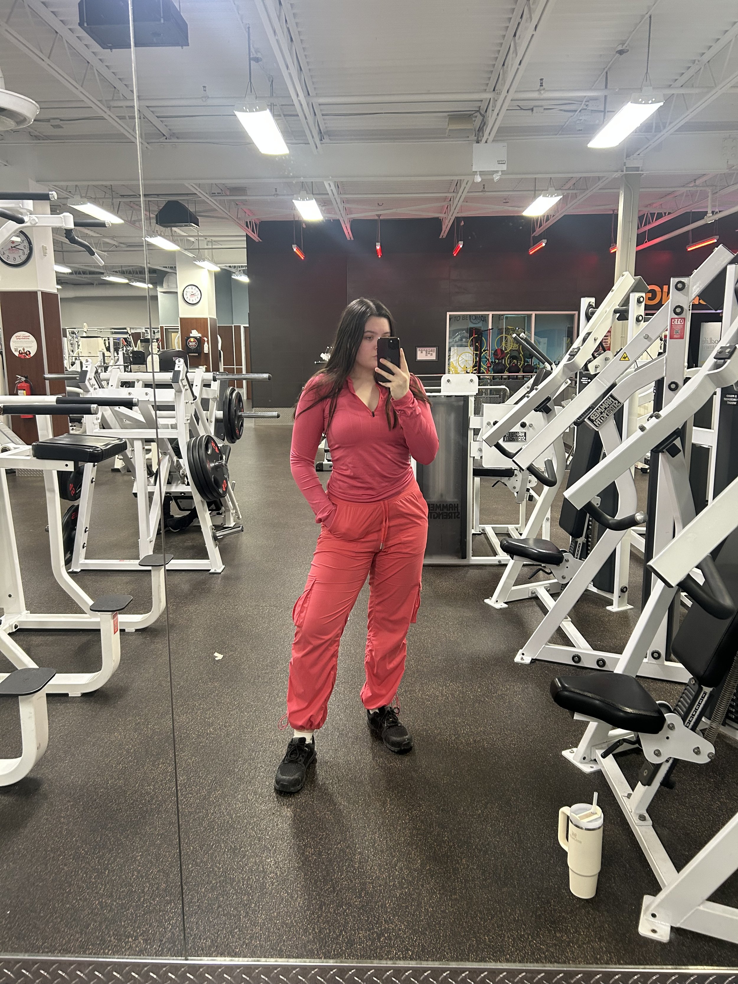 Abi Mack wearing a pink athletic tracksuit and black sneakers taking a selfie in a gym mirror surrounded by exercise equipment.
