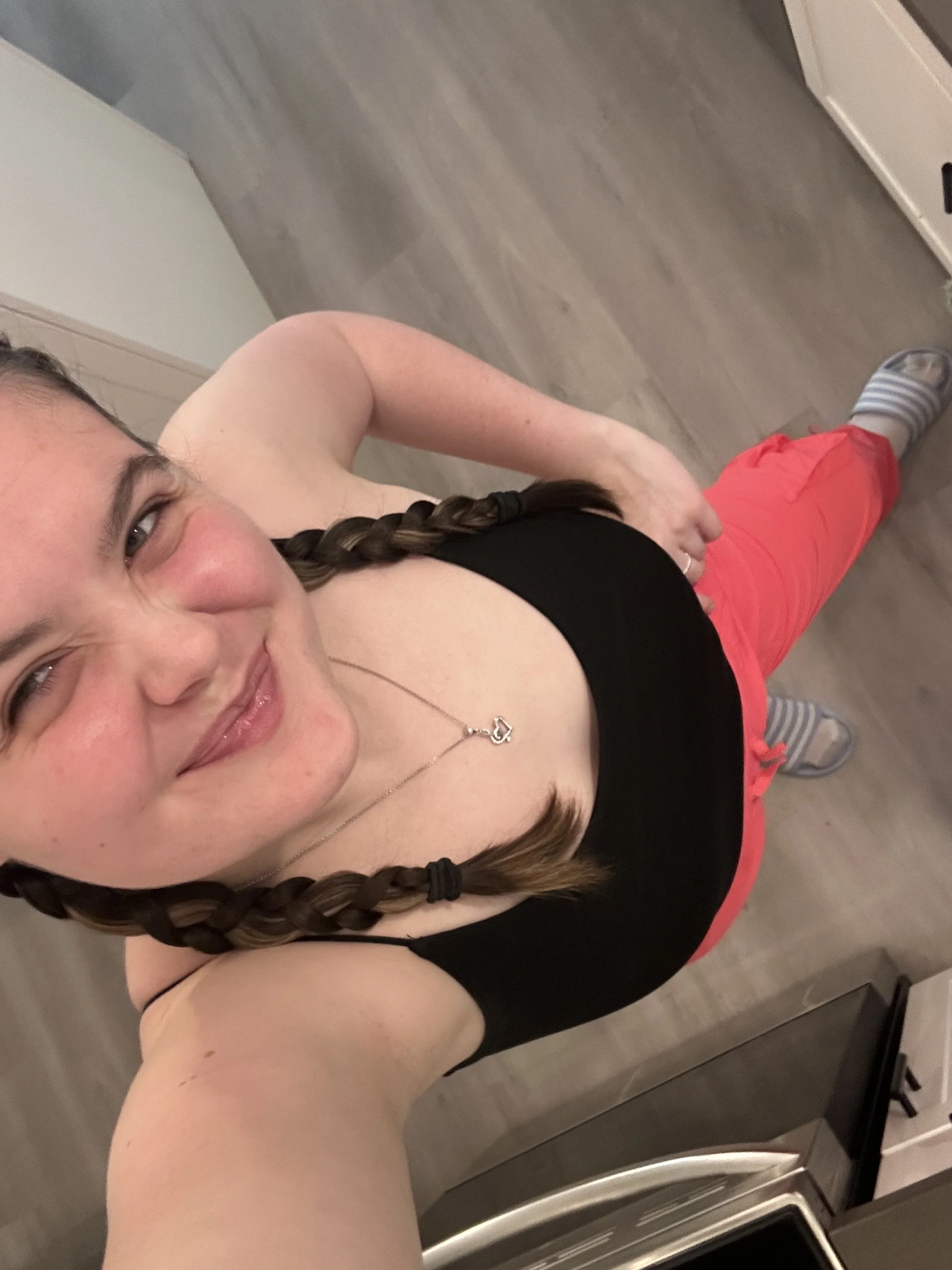 Abi Mack with braided hair taking a selfie from above, smiling, wearing a black top, pink pants, and striped socks, standing in a kitchen with a gray floor and white cabinets.