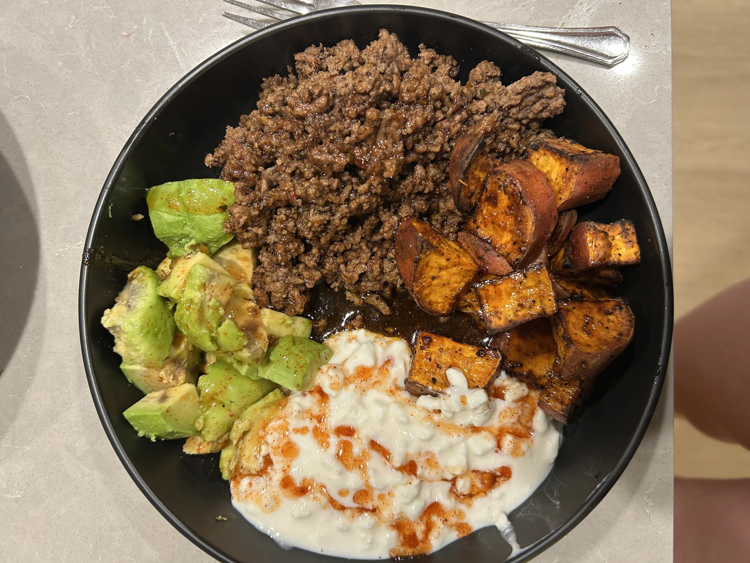 A bowl containing cooked ground beef, roasted sweet potatoes, sliced avocado with seasoning, and sour cream garnished with hot sauce.