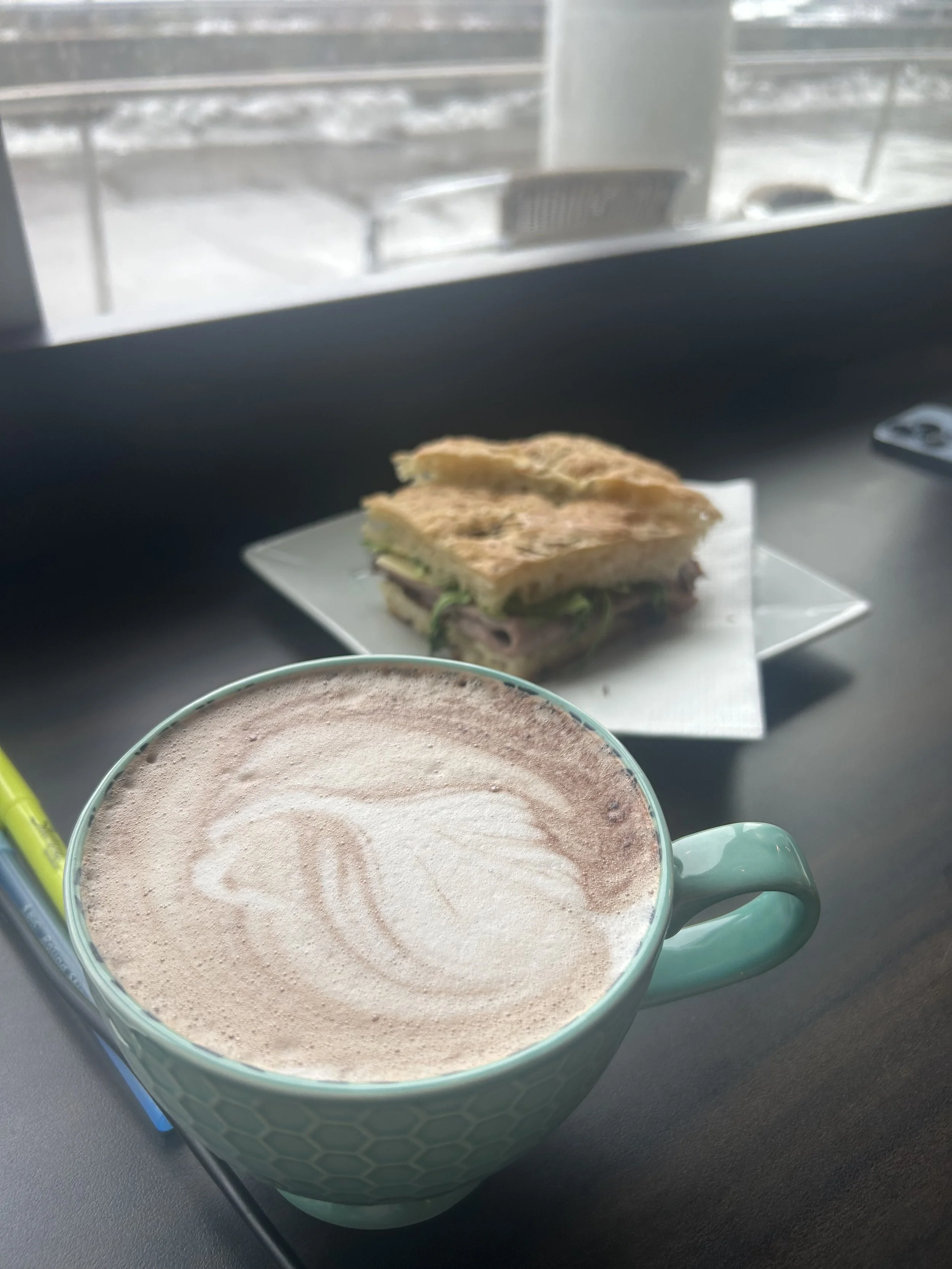 A mug of frothy hot chocolate and a sandwich on a plate are on a black table near a window with a view of snow outside.