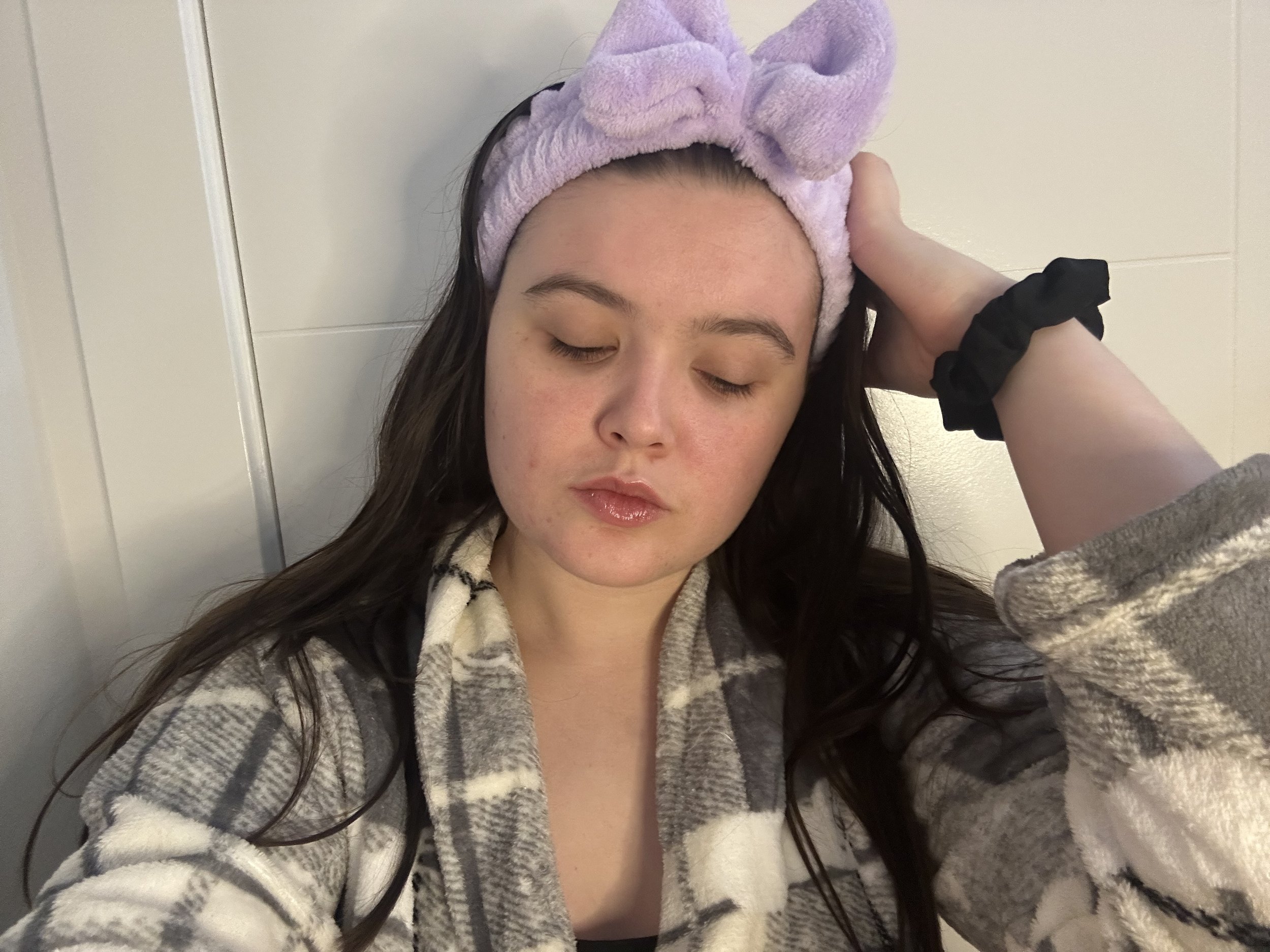 A woman with long dark hair, closed eyes, shiny lips, wearing a purple headband with a bow and a fleece jacket, leaning against a white wall with her hand on her head.