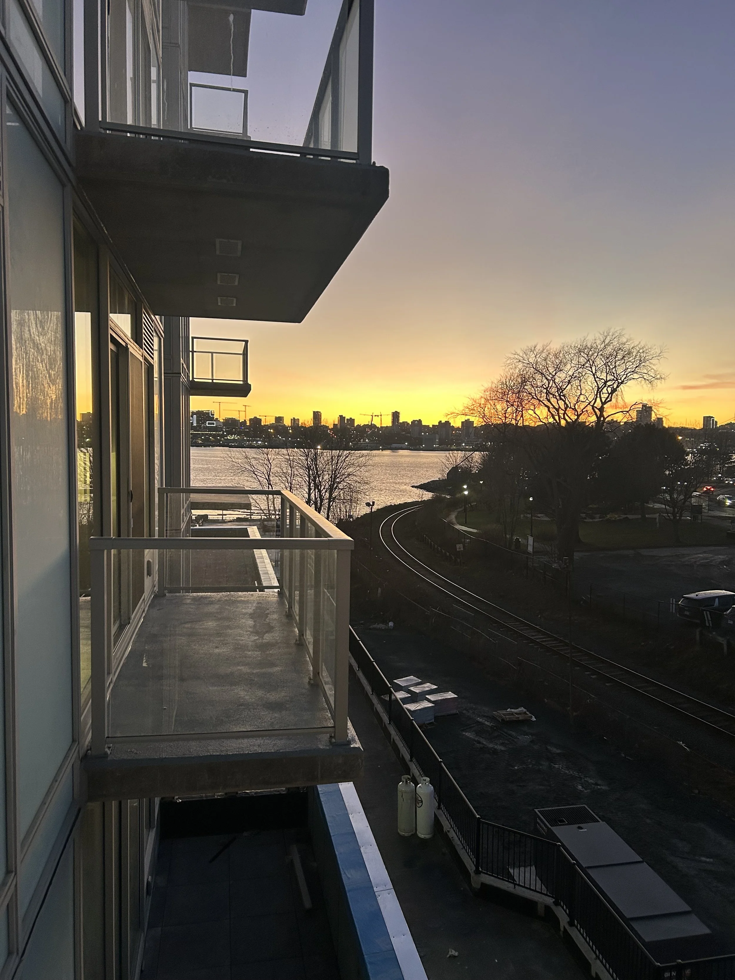 Sunset view from a balcony overlooking a river, with city skyline in the distance, and bare trees in the foreground.