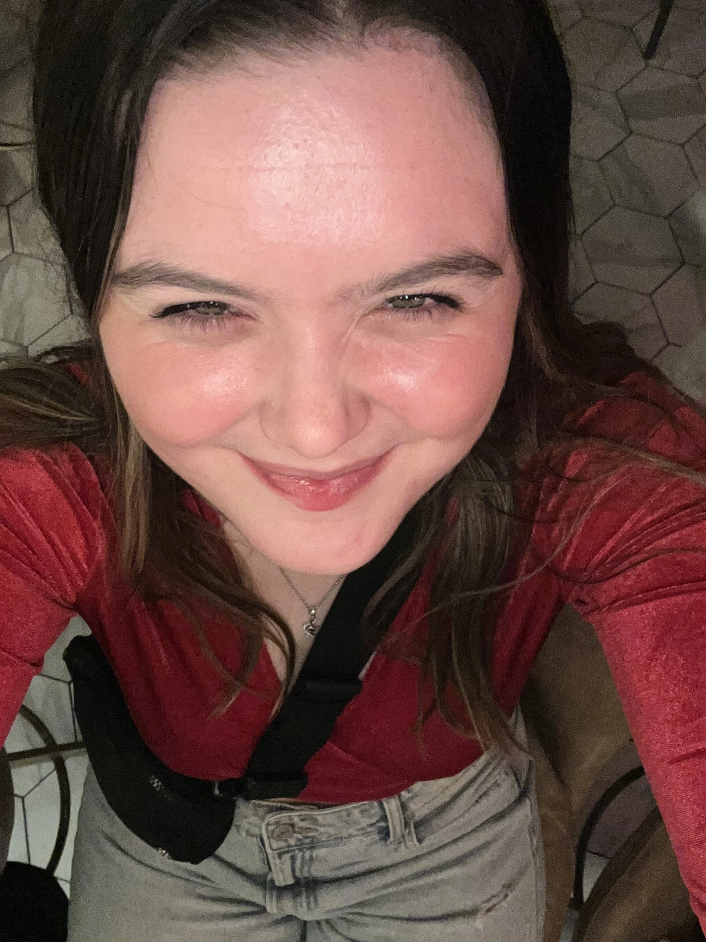 A woman taking a selfie from above, smiling with her eyes slightly squinted, wearing a red top and light-colored jeans, standing on a hexagonal-tiled floor.