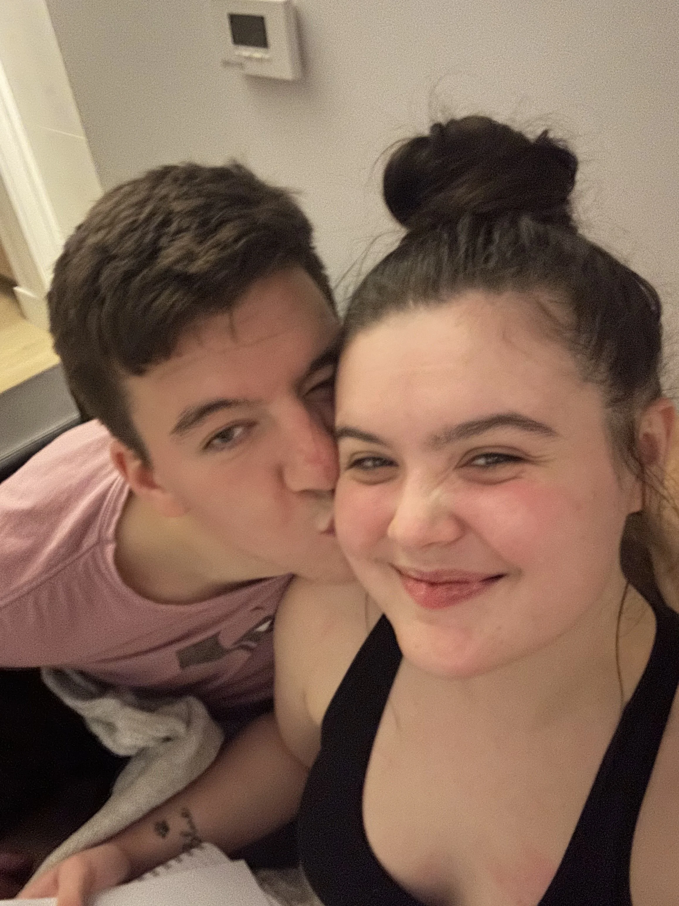 A young man is kissing Abi Mack on the cheek while she smiles with her eyes closed, in a close-up selfie.