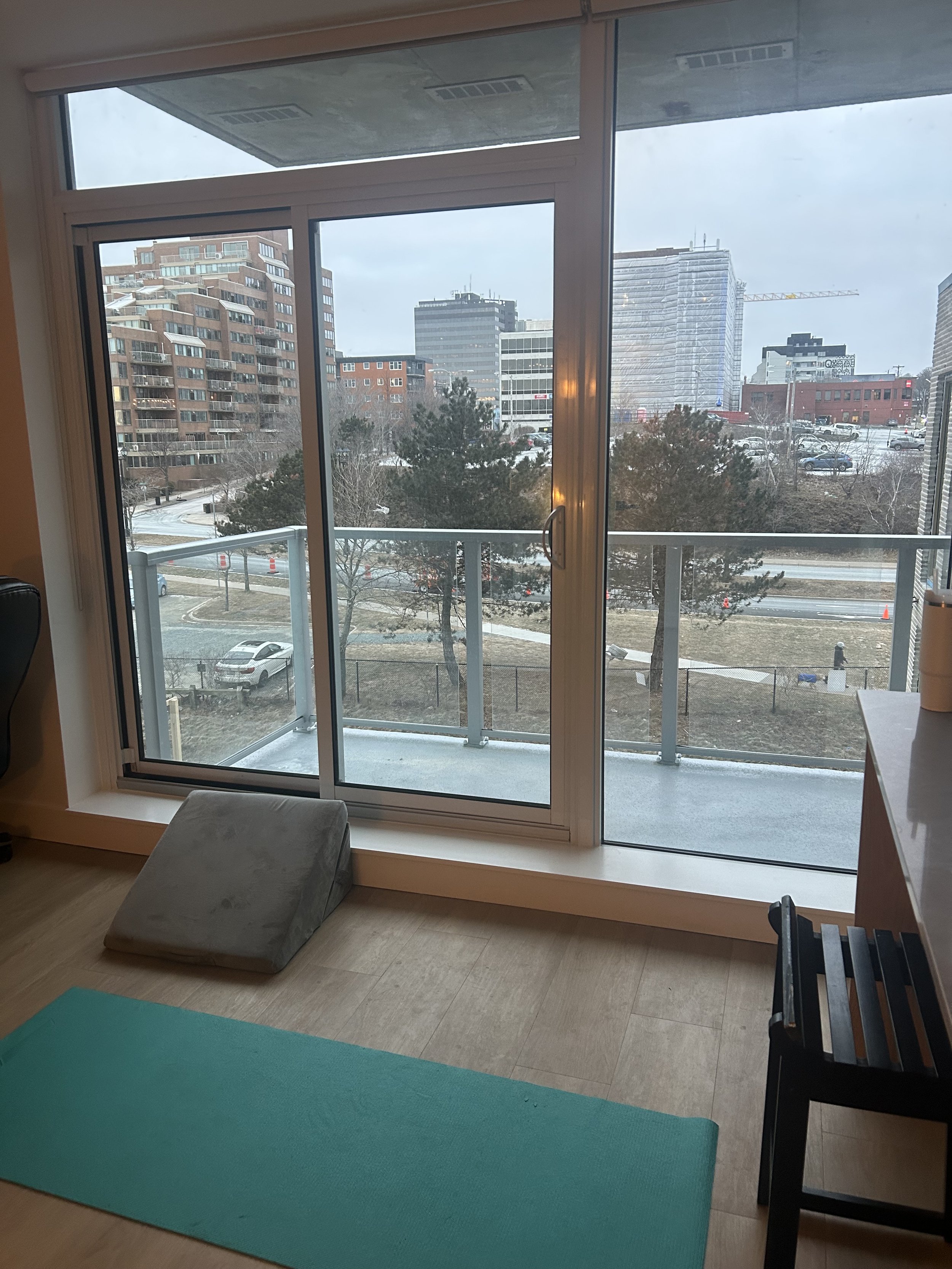 A room with a large glass door leading to a balcony, overlooking a cityscape with tall buildings, trees, and a parking lot. There is a gray cushion on the wooden floor and a teal yoga mat.
