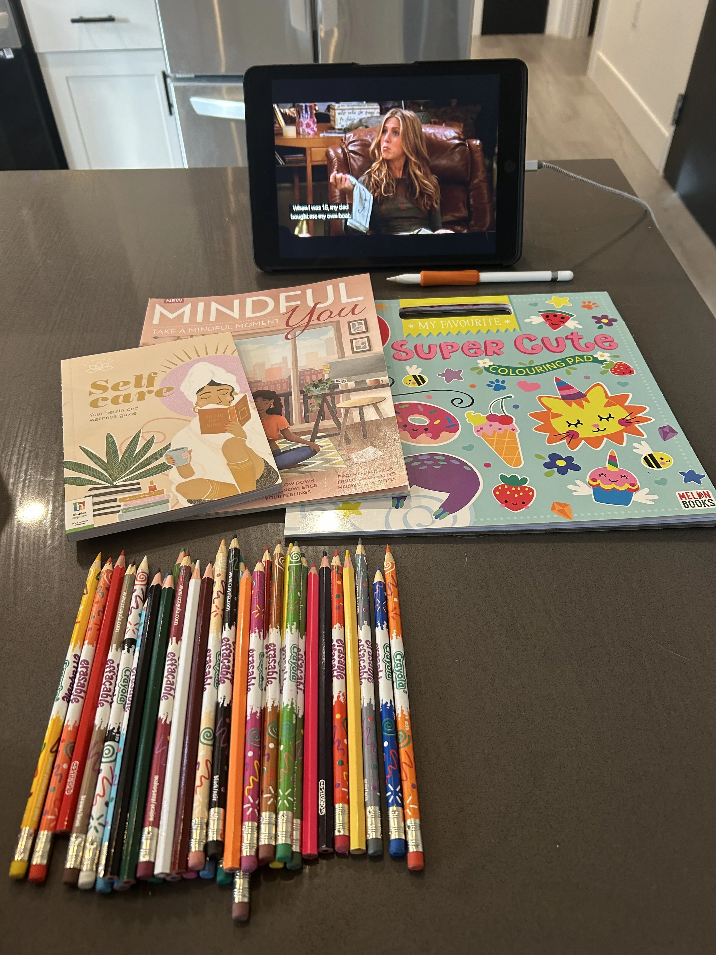 A tablet on a table displaying a scene from a TV show with a woman sitting on a couch. In front of the tablet are colorful books, a packet of coloring pencils, a coloring pad, and a pen.
