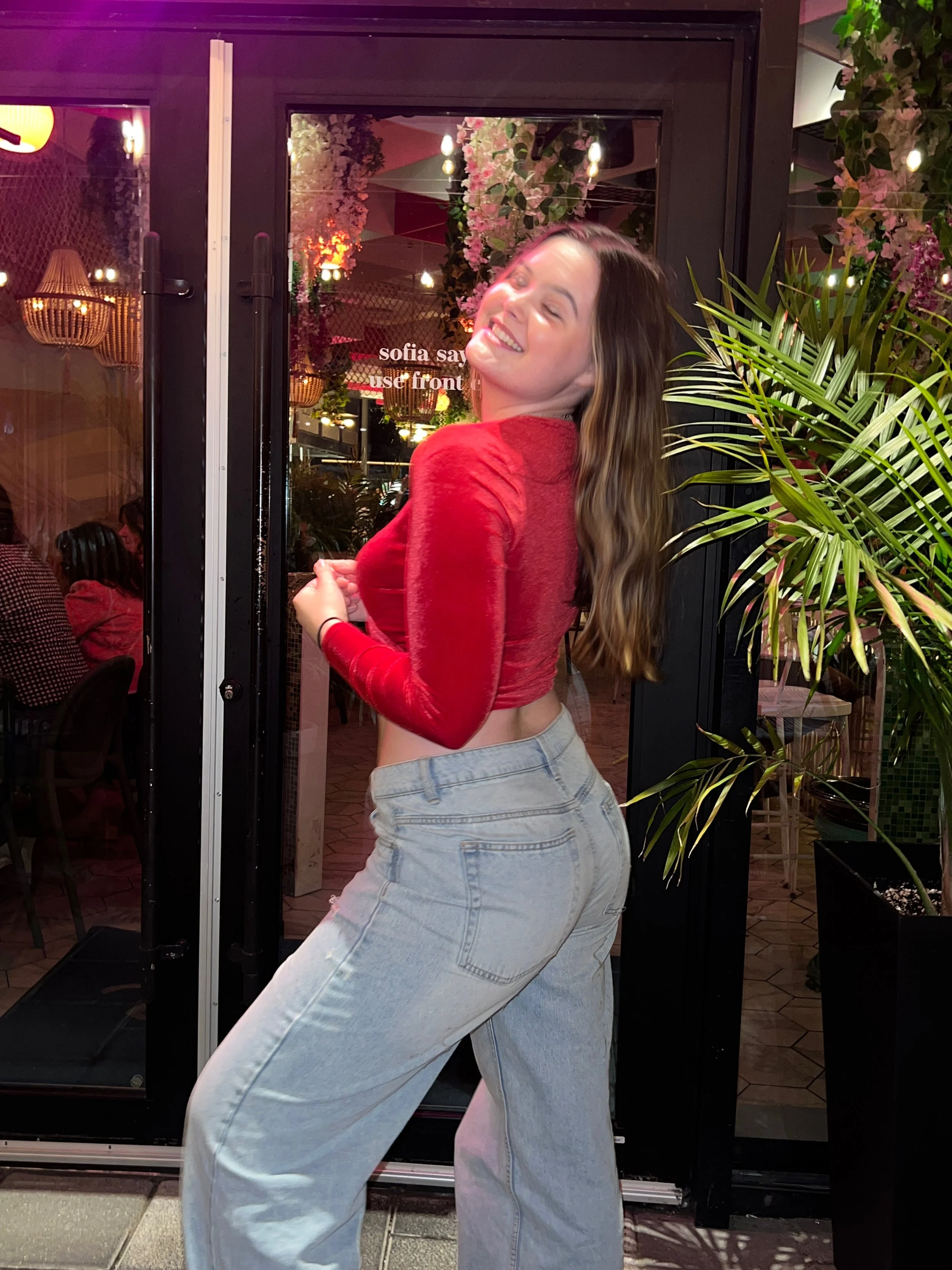 A woman with long brown hair smiling and posing with her eyes closed, wearing a red long-sleeve crop top and light blue oversized jeans, standing outside a restaurant with indoor plants and pink flowers visible through a glass door.