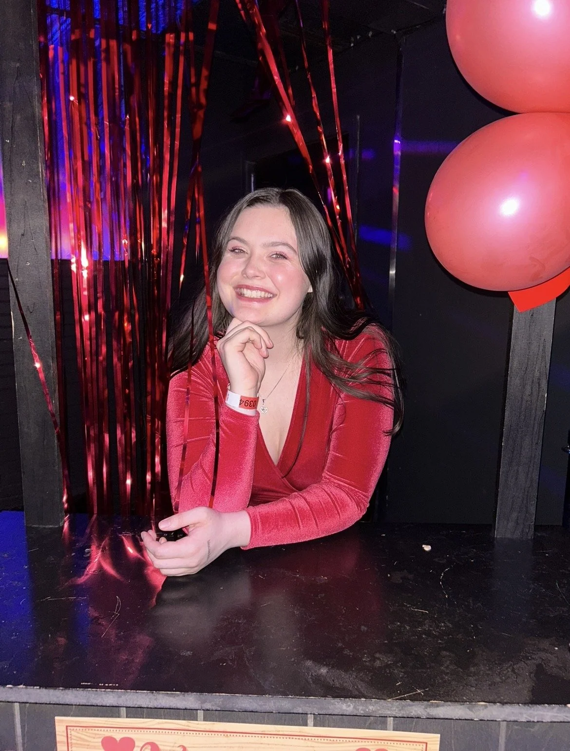 A young woman with long dark hair smiling, dressed in a red long sleeve top, sitting at a decorated booth with red streamers and pink balloons, in a festive setting.
