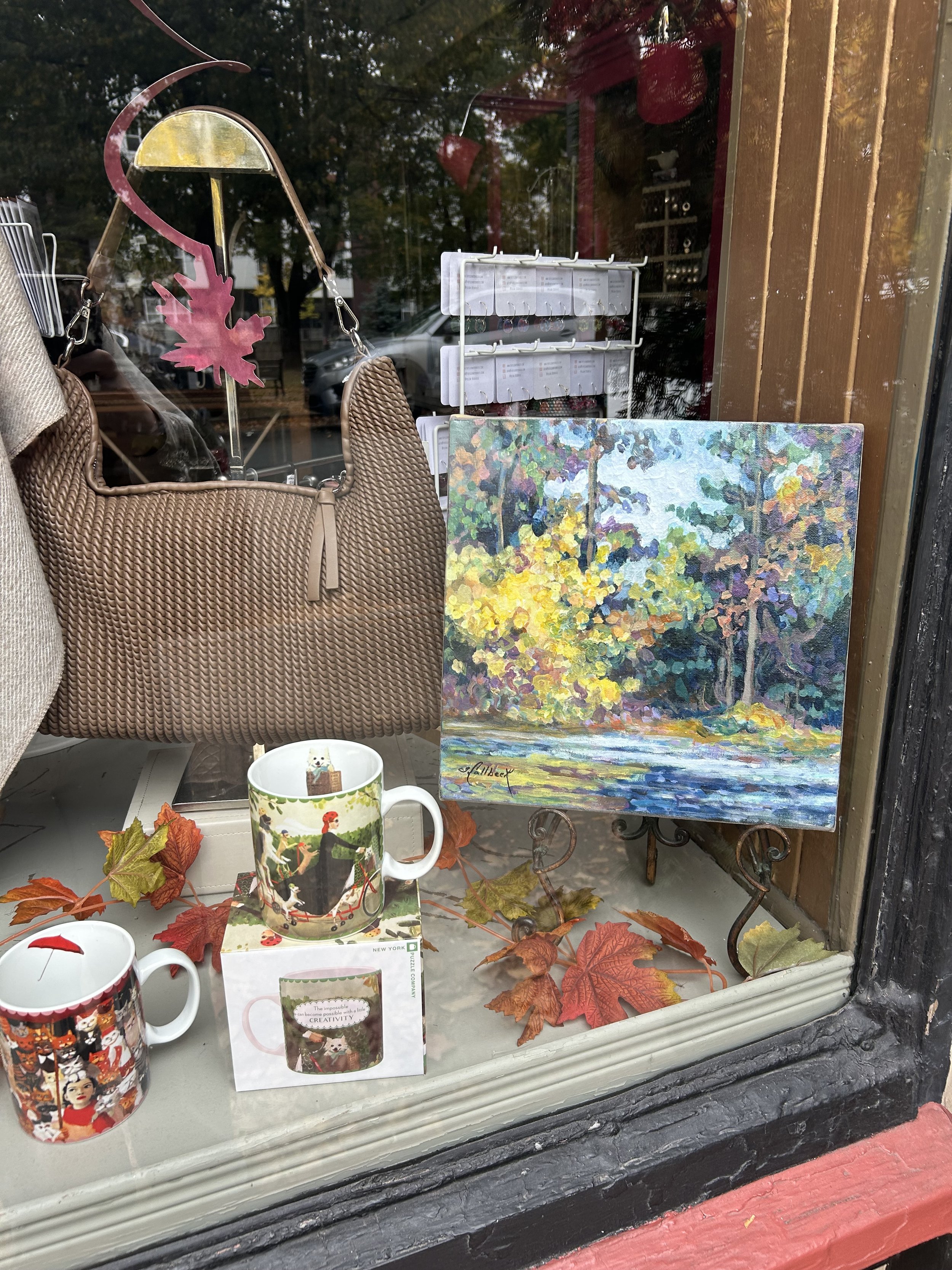 A window display with a brown purse, two decorative mugs, a small colorful painting of a river and trees, autumn leaves, and a white plastic rack with small items inside.