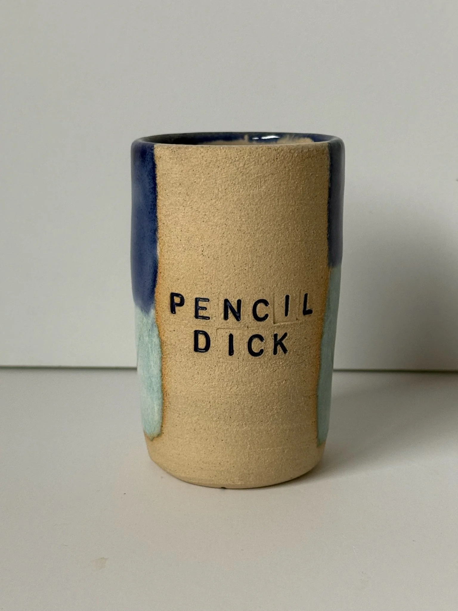 A ceramic mug with the words 'PENCIL DICK' embossed on the front, featuring a textured beige surface and blue and green glazing accents.