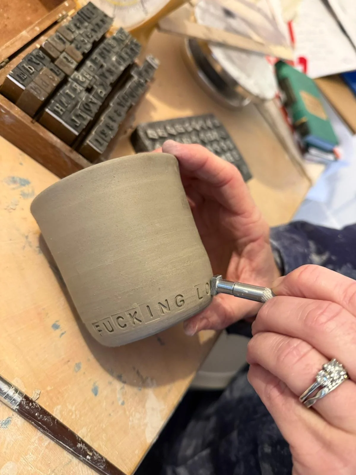 A person is carving words into a ceramic mug, which reads 'FUCKING LOVE'. The person's hand is holding the mug steady while using a tool to inscribe the text.