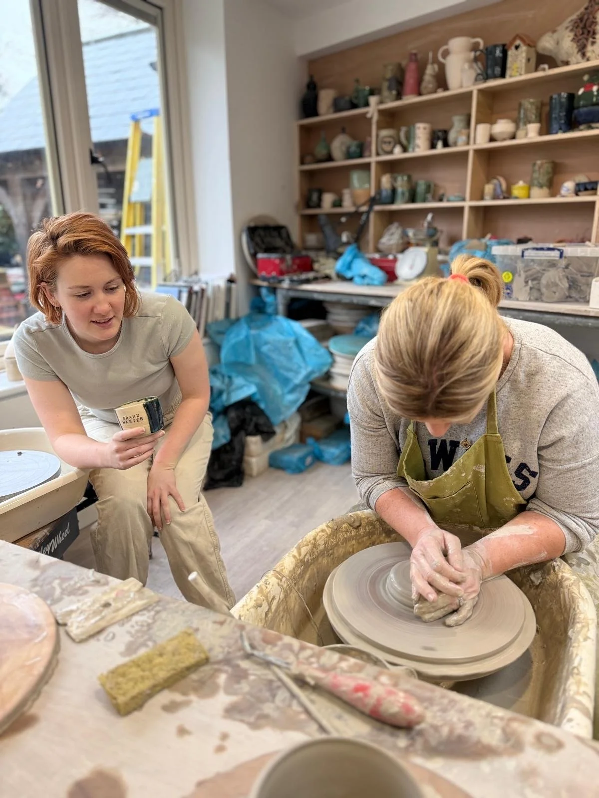 Two women working on a pottery wheel in a ceramic studio, with shelves of pottery and tools in the background, and one woman holding a ceramic piece while the other shapes clay on the wheel.