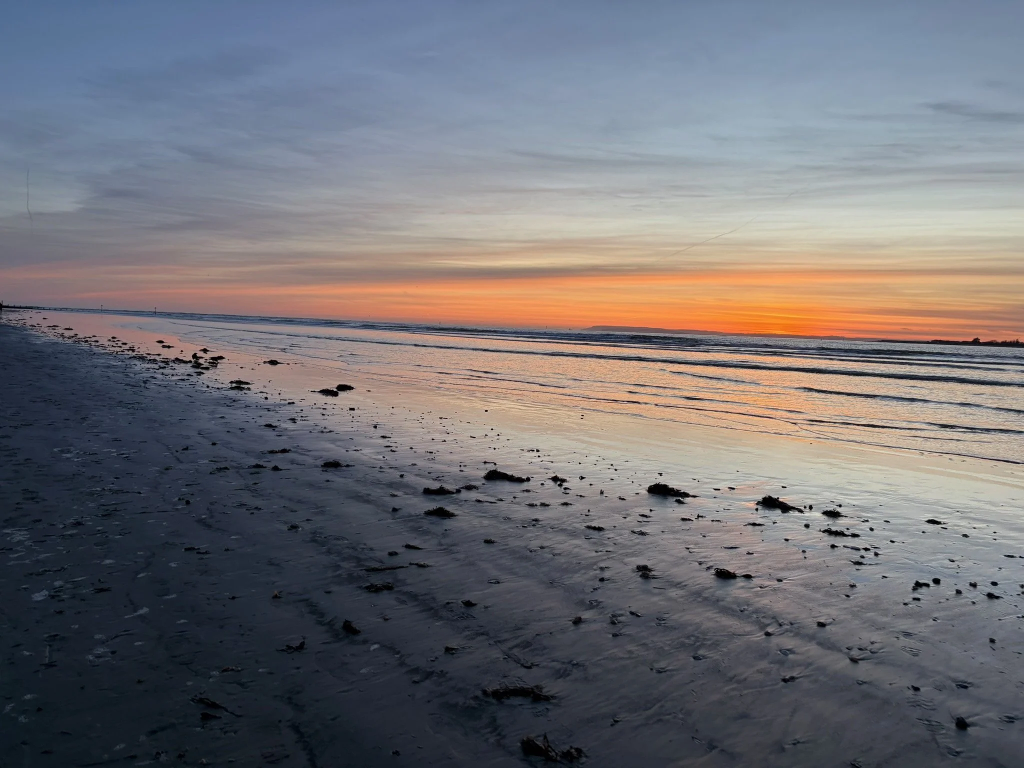 Sunset over the beach with calm waves, wet sand, and scattered seaweed and rocks.