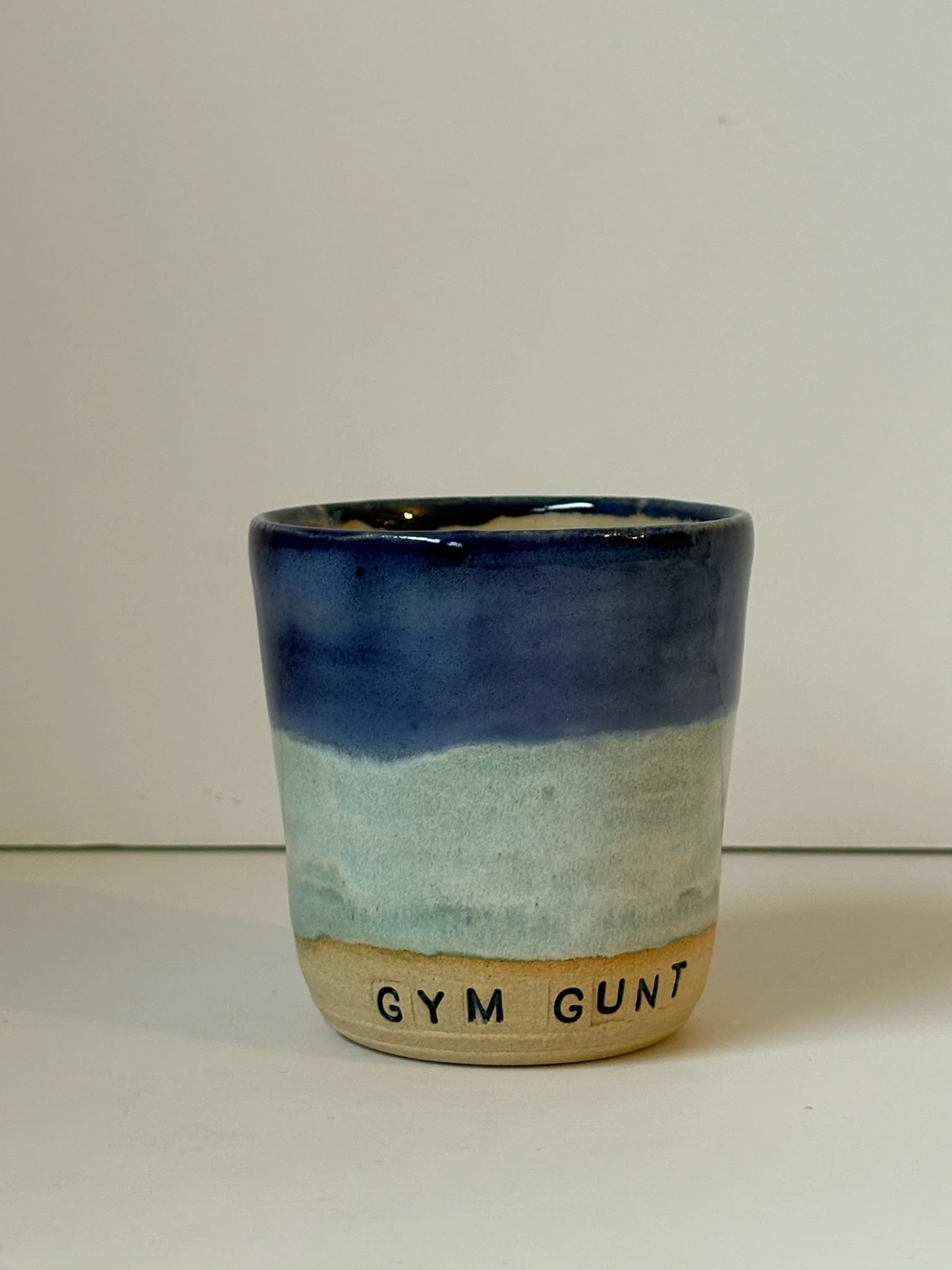 Ceramic mug with a color gradient from beige to blue with the words 'GYM GUNT' printed at the bottom.