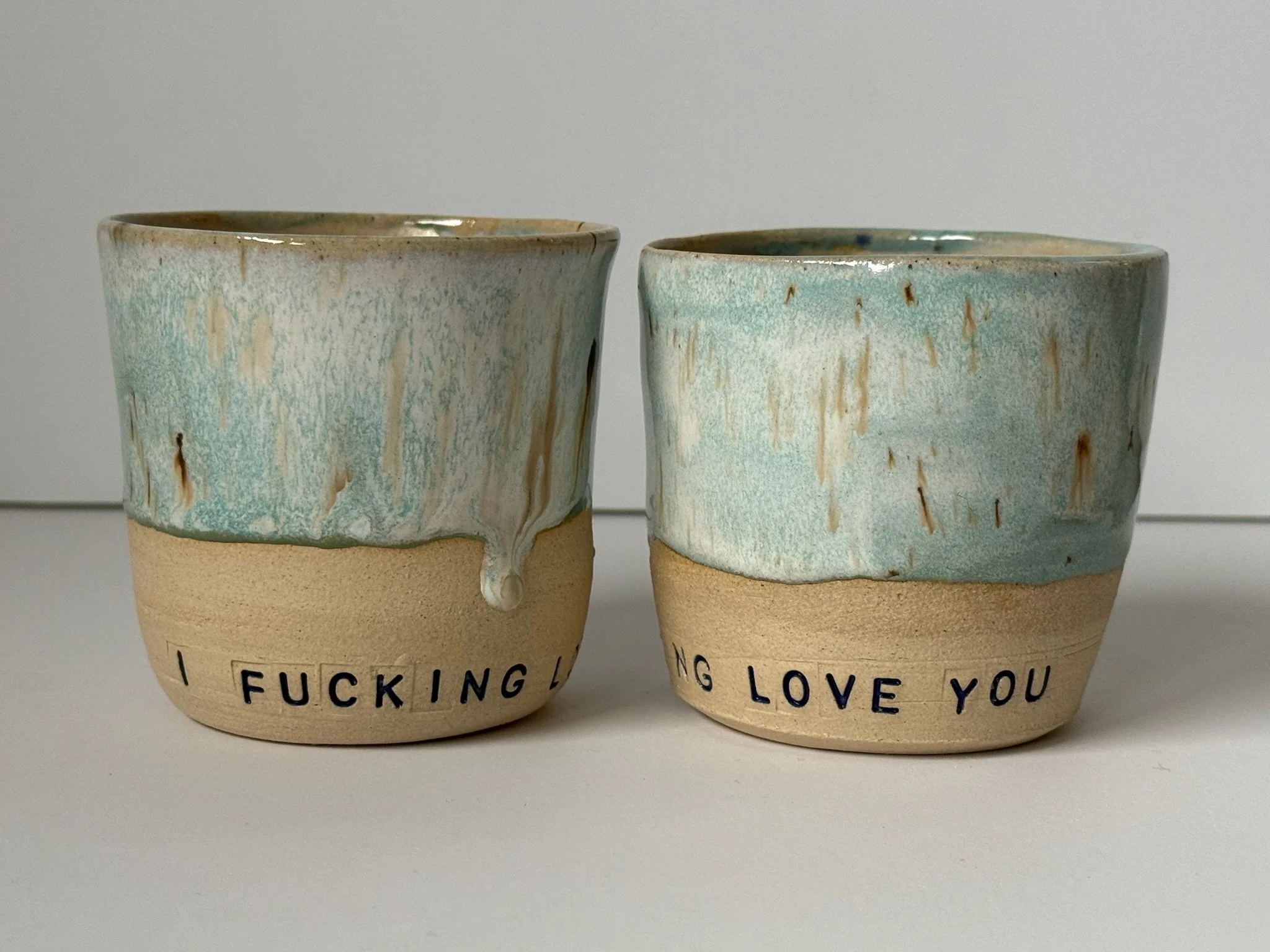 Two ceramic cups with blue and beige glaze, one displaying partial text 'I FUCKING L...' and the other showing 'NG LOVE YOU'.