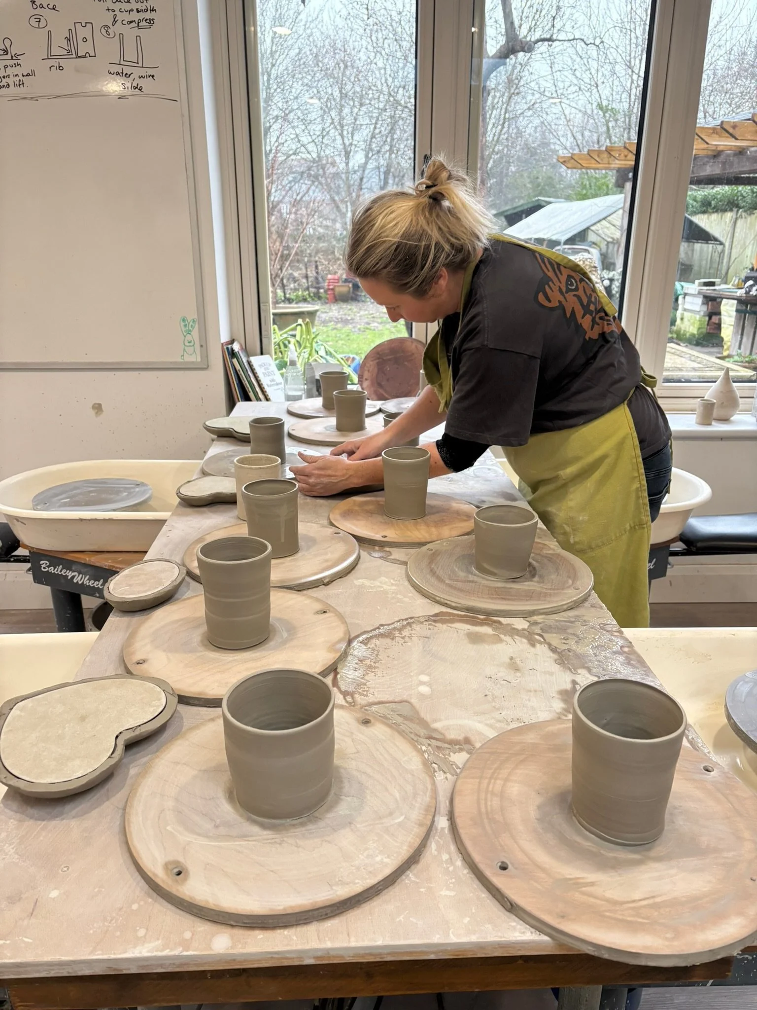 A woman in a black shirt and green apron working on pottery, arranging clay cups on wooden bases in a pottery studio with large windows, outside trees, and garden visible.