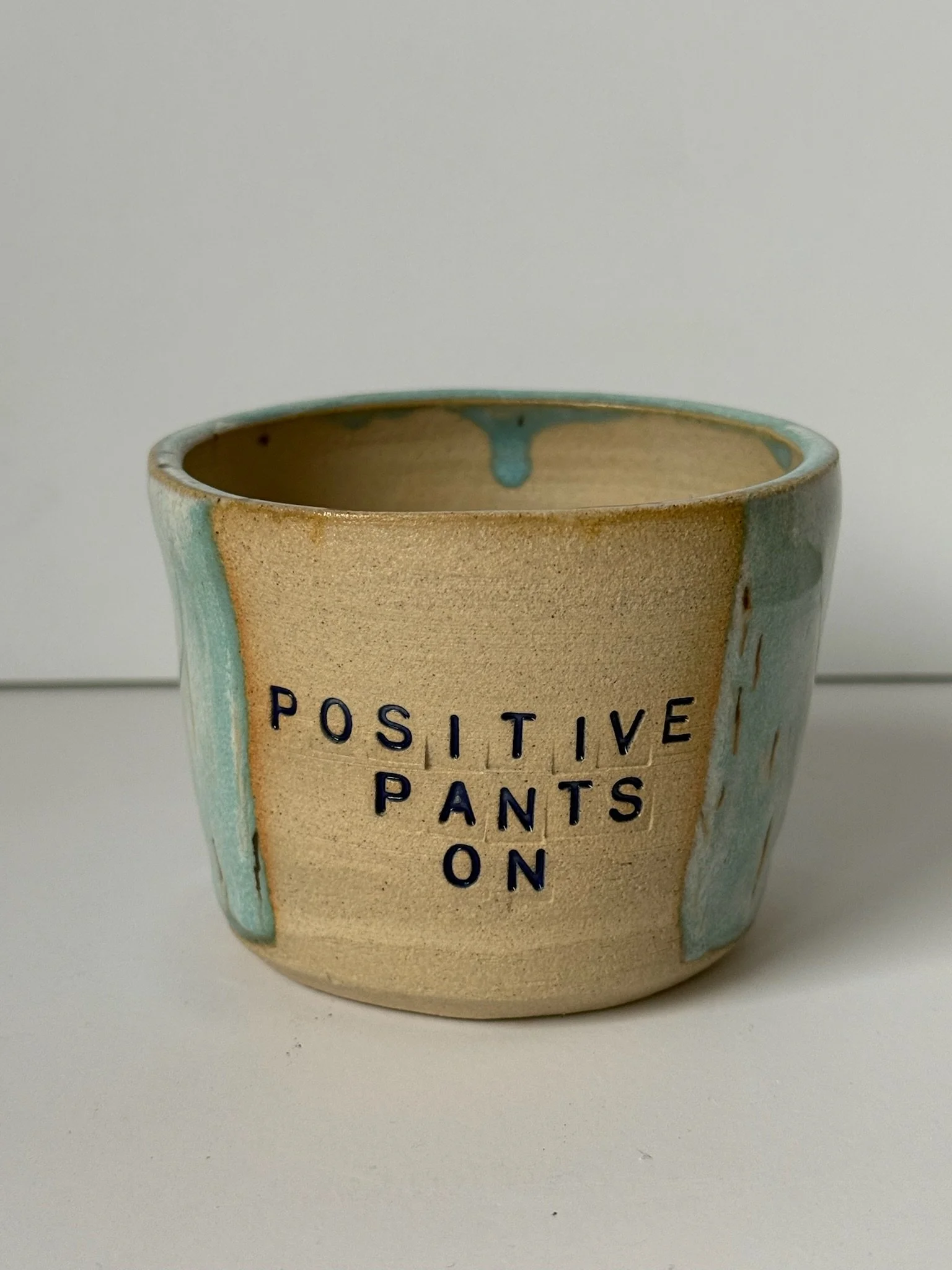 A ceramic pot with pastel blue and beige glaze, featuring the phrase 'POSITIVE PANTS ON' in black lettering.