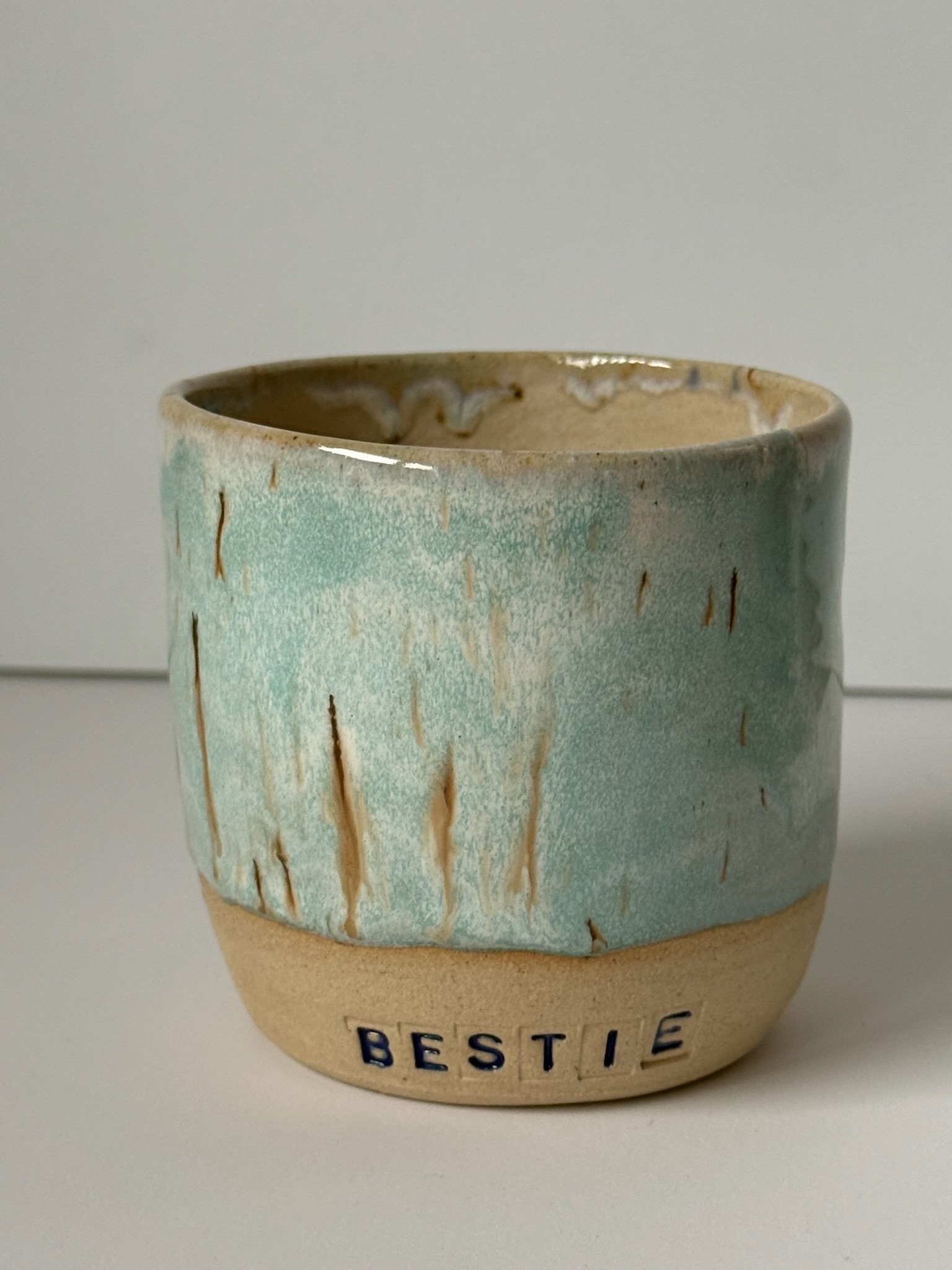 A ceramic mug with a beige bottom and a light blue, textured glaze on the upper part, with the word 'BESTIE' in blue letters on the bottom edge.