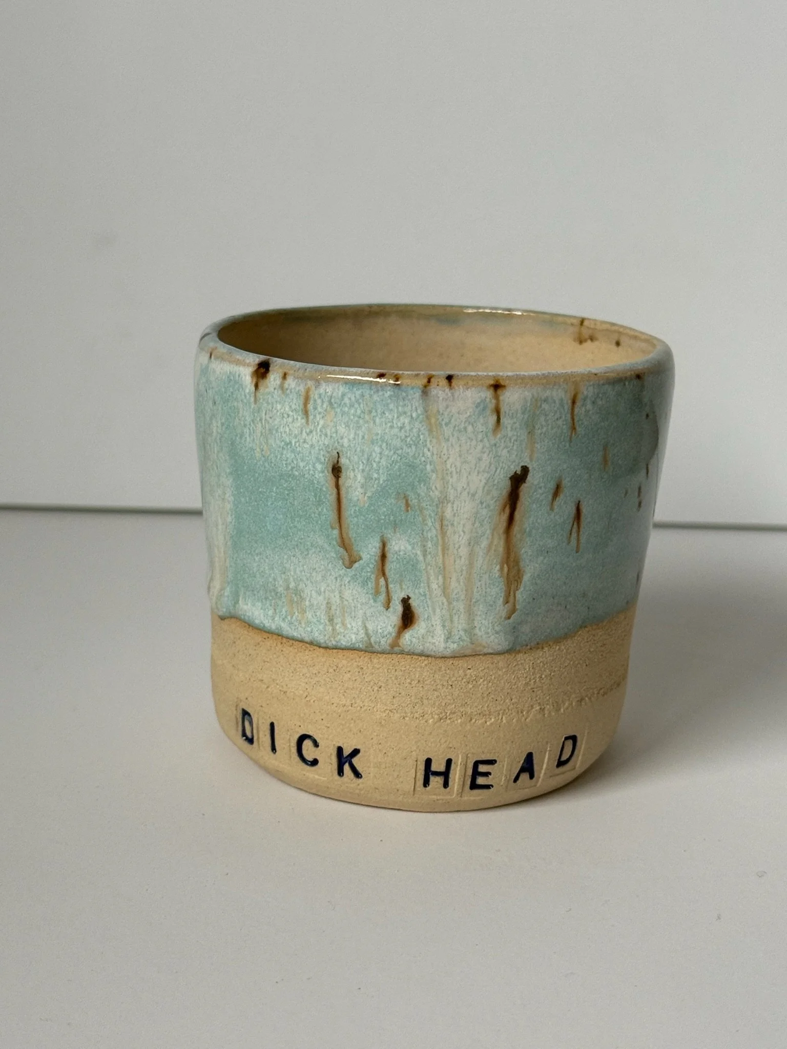 A ceramic mug with a blue and brown glaze, with the words 'DICK HEAD' written on the lower part.