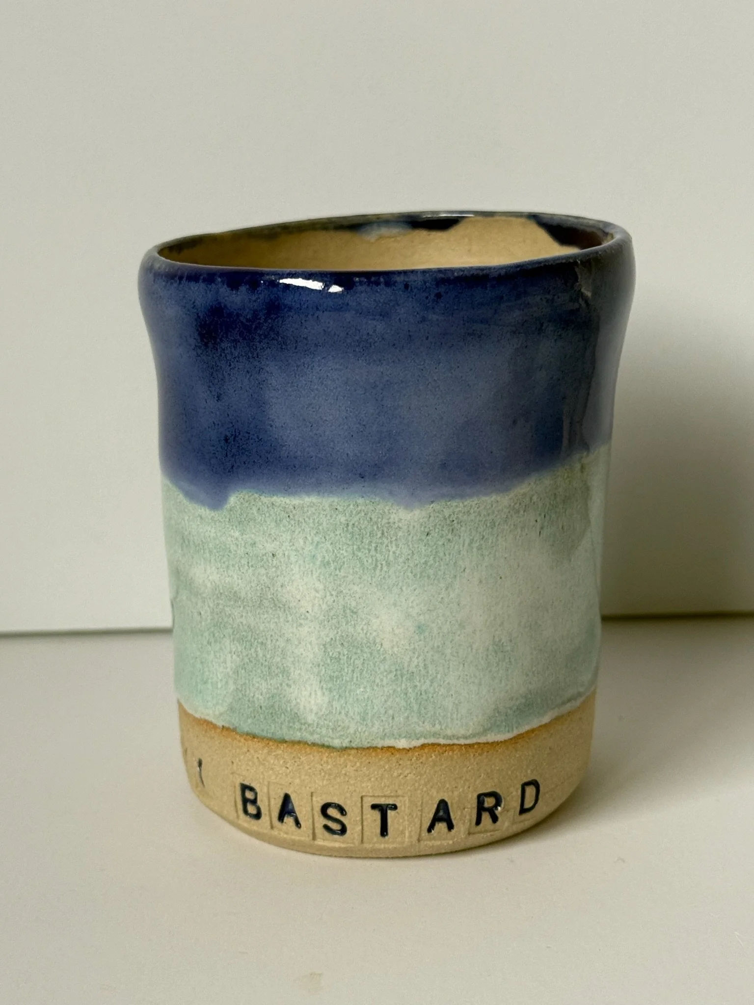 A ceramic mug with blue, green, and beige glaze, with the word 'BASTARD' spelled out in letter tiles at the bottom.