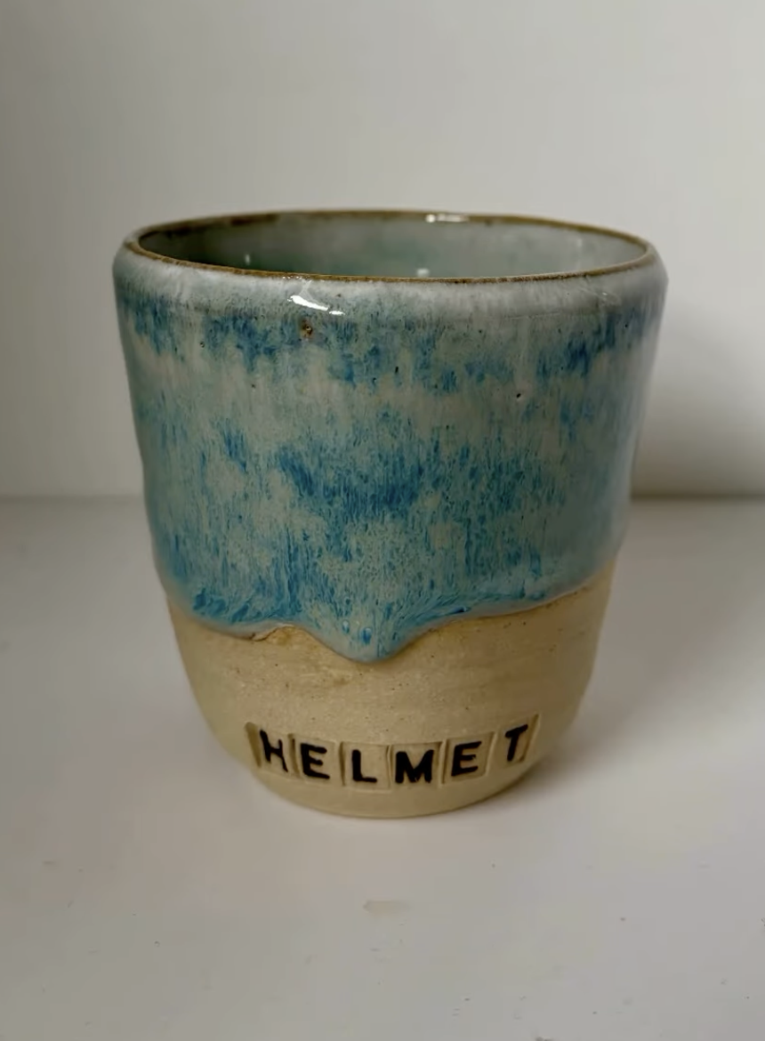 A ceramic mug with a blue glazed top and a beige bottom, with the word 'HELMET' spelled out in black letter tiles on the front.