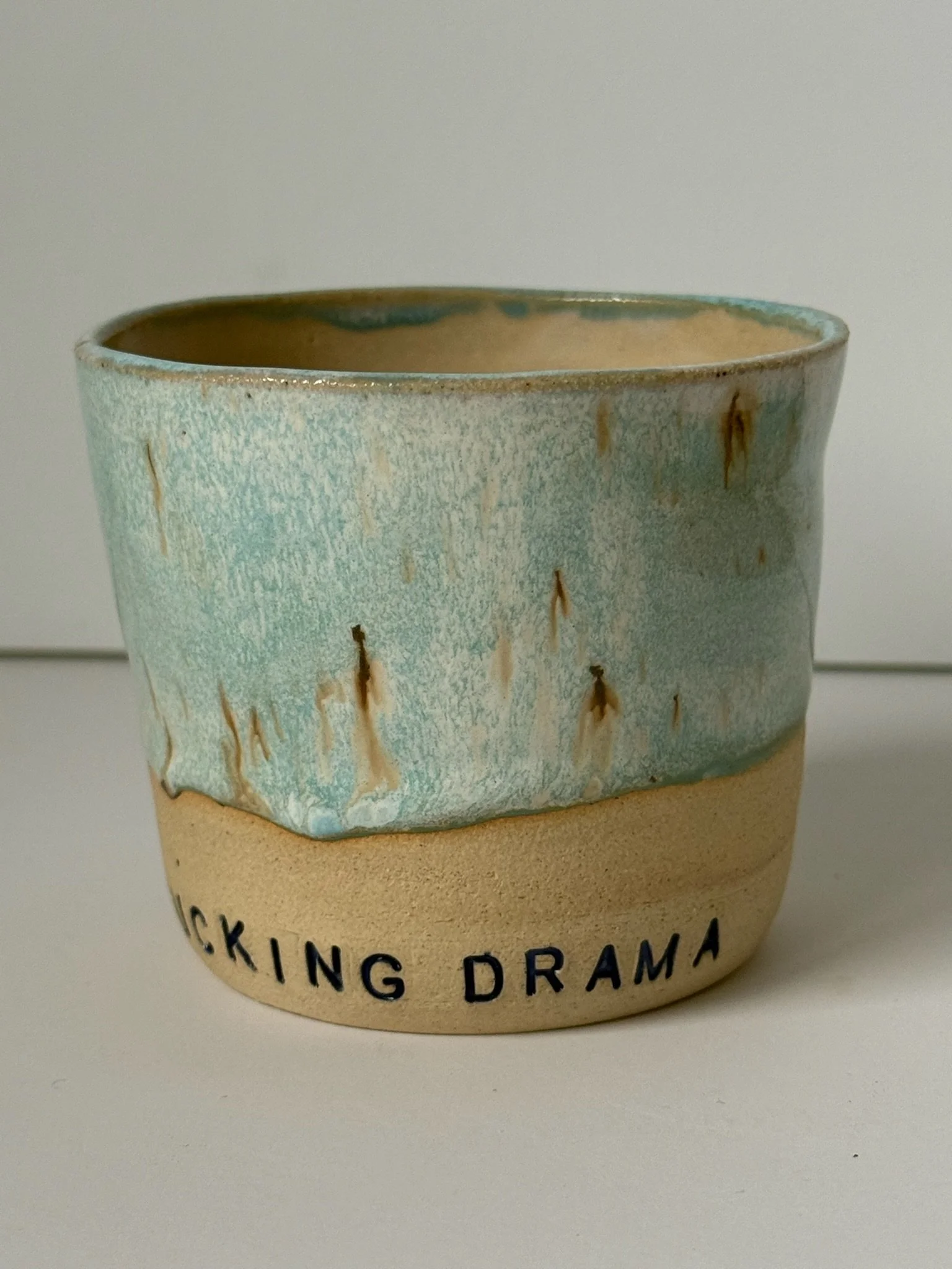 Ceramic mug with a glazed pale blue top half and a tan bottom half, featuring the text 'LICKING DRAMA' on the bottom.