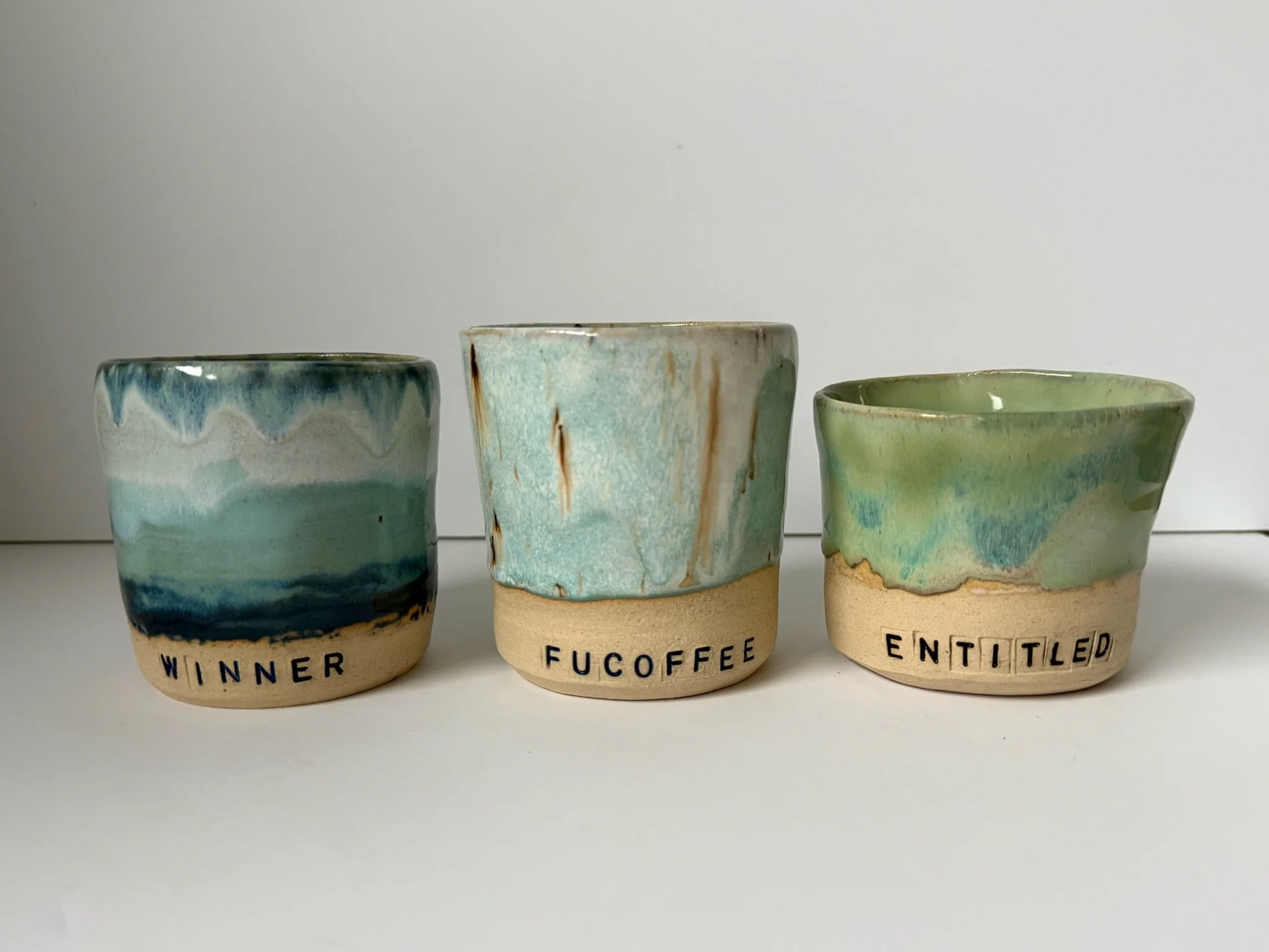 Three ceramic cups with watercolor-like glaze and handwritten labels: 'WINNER', 'FUCOFFEE', and 'ENTITLED'.