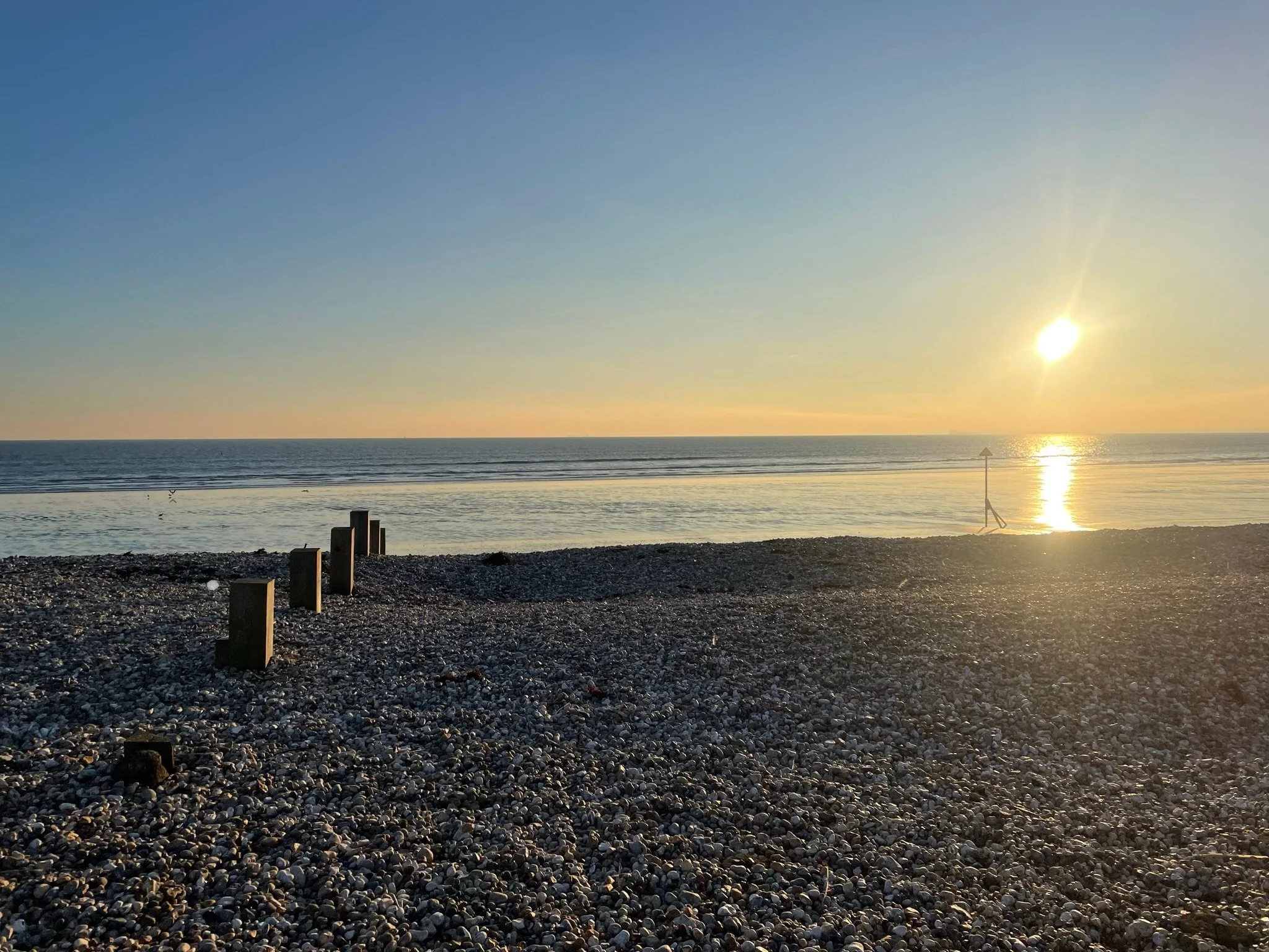 Sunset over a pebble beach with wooden posts and a solitary navigational sign by the water.