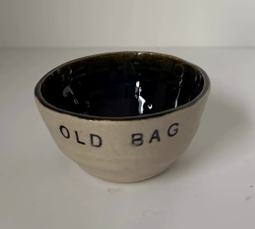 A small ceramic bowl with black interior and beige exterior, with the words 'OLD BAG' written on it, placed on a white surface.