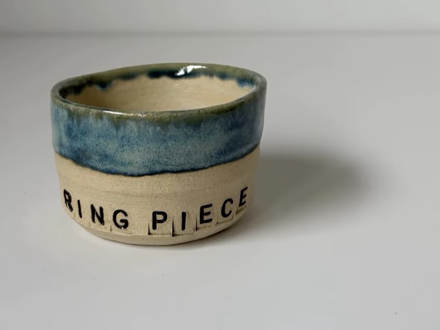 A small ceramic bowl with a beige exterior and blue glaze inside, featuring the words 'RING PIECE' spelled out with letter beads or blocks on the side.