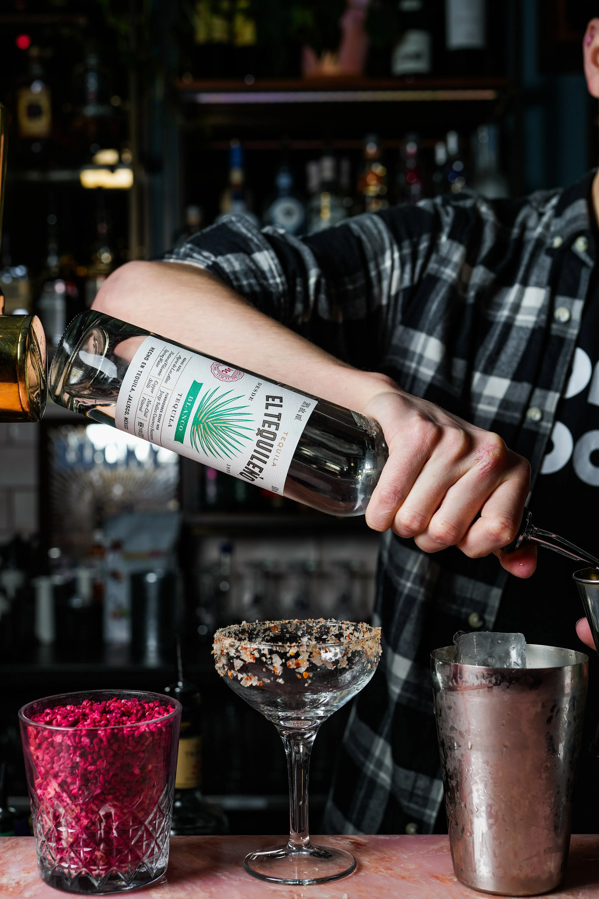 Bartender in plaid shirt pouring tequila into a glass with a salted rim, on a bar counter with pink garnish and ice bucket nearby.