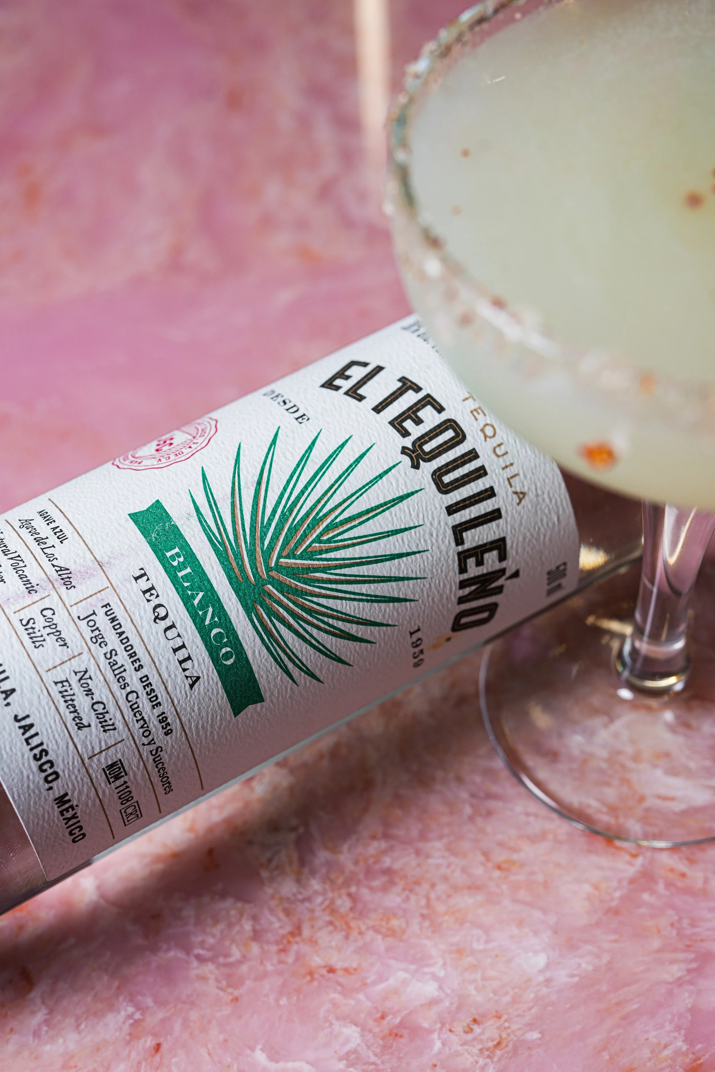A bottle of El Tequileño Blanco tequila lying on a pinkish-orange textured surface, next to a glass filled with margarita.