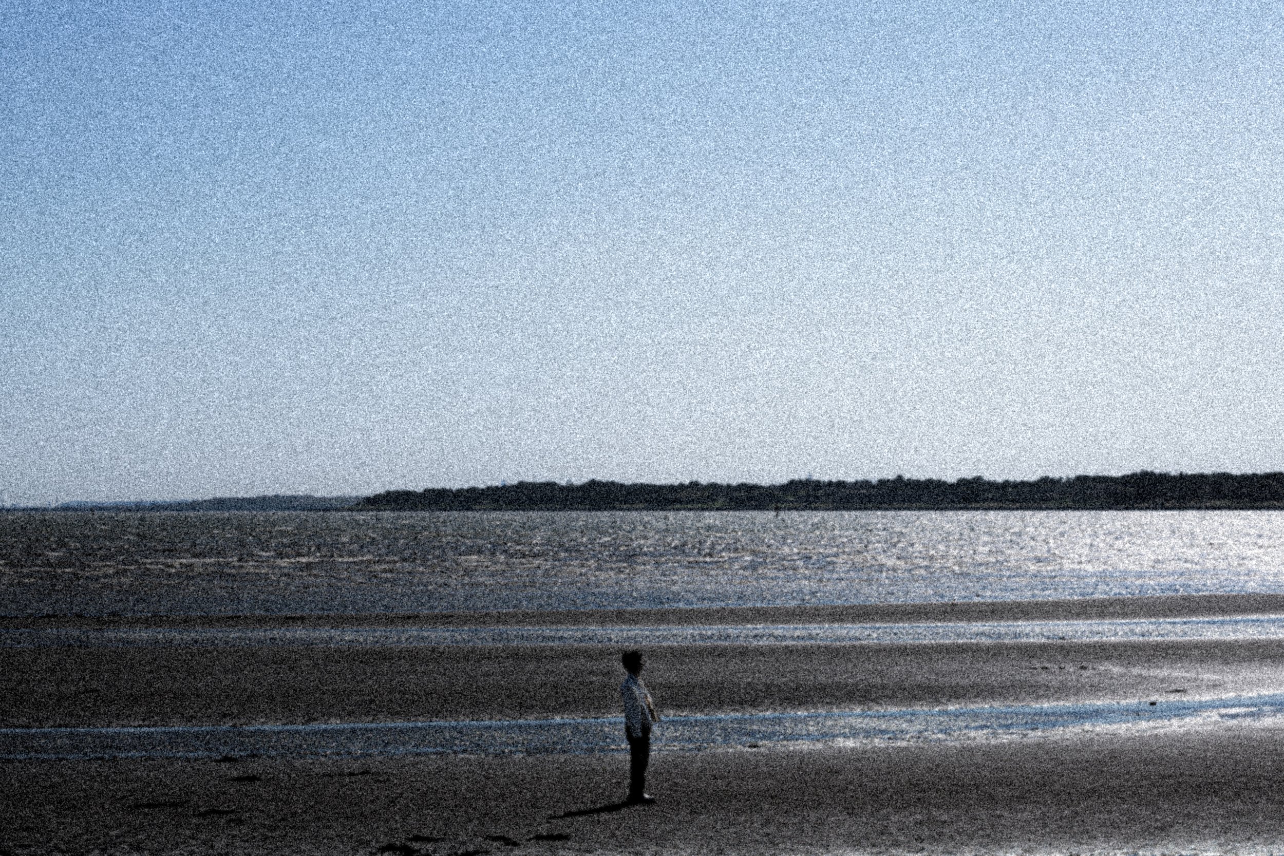 海辺に一人の子供が立っている風景。夕暮れ時で、静かな海と空、遠くに島のような陸地が見える。