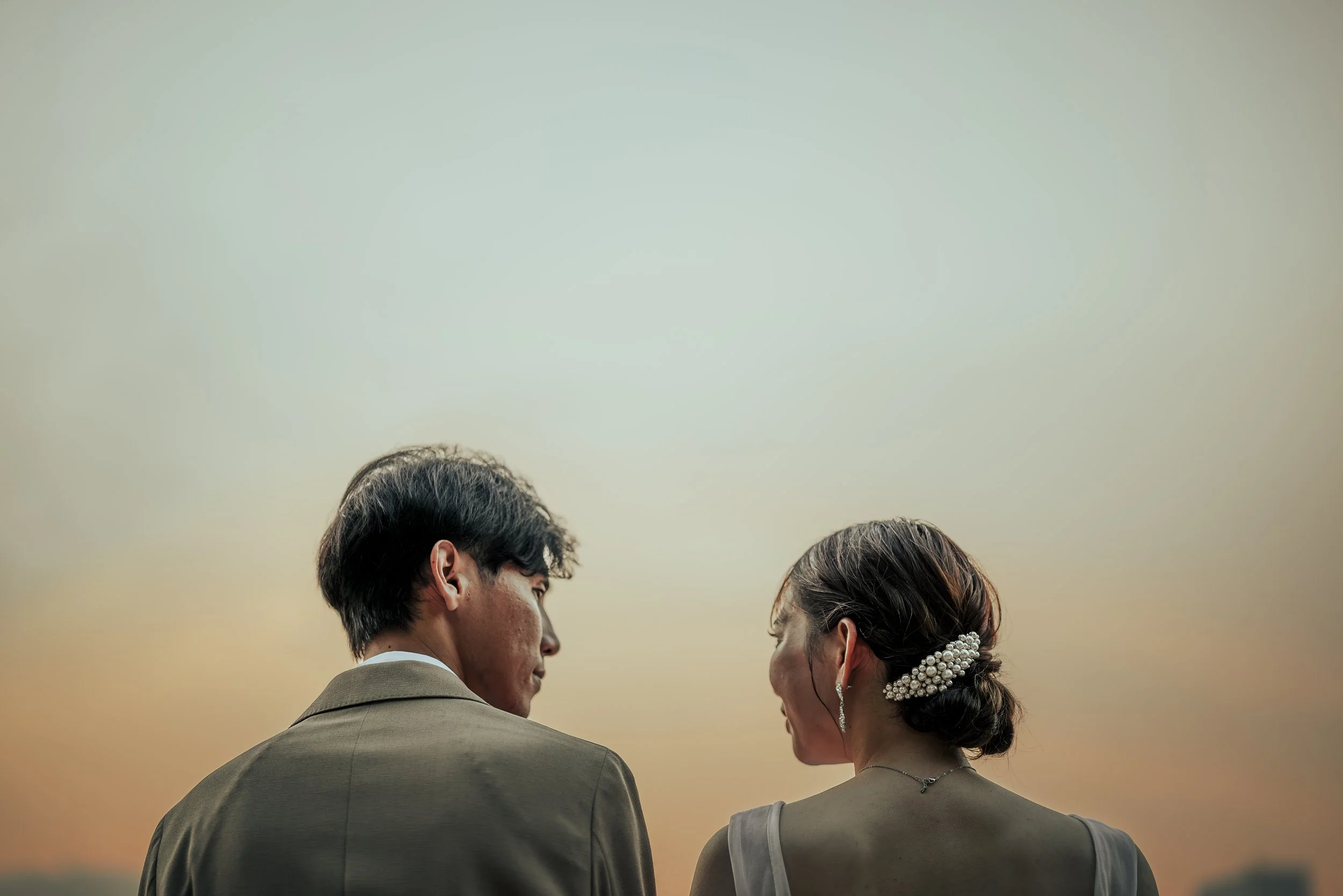 夕焼けの空の下、結婚式のドレスを着た女性とスーツを着た男性が横顔で向き合っている場面