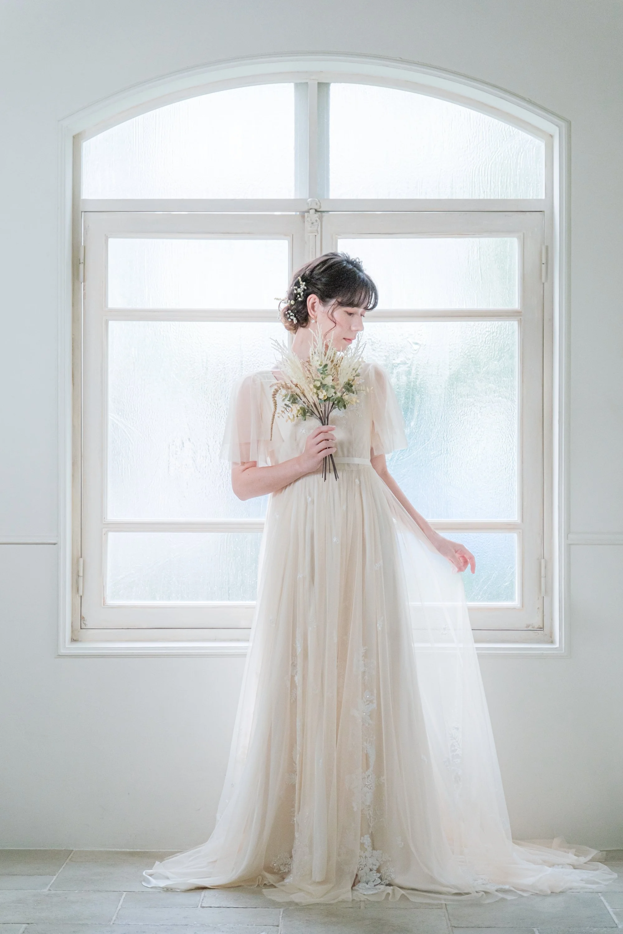 白いドレスを着た女性が窓の隣で花束を持ちながら微笑んでいる写真