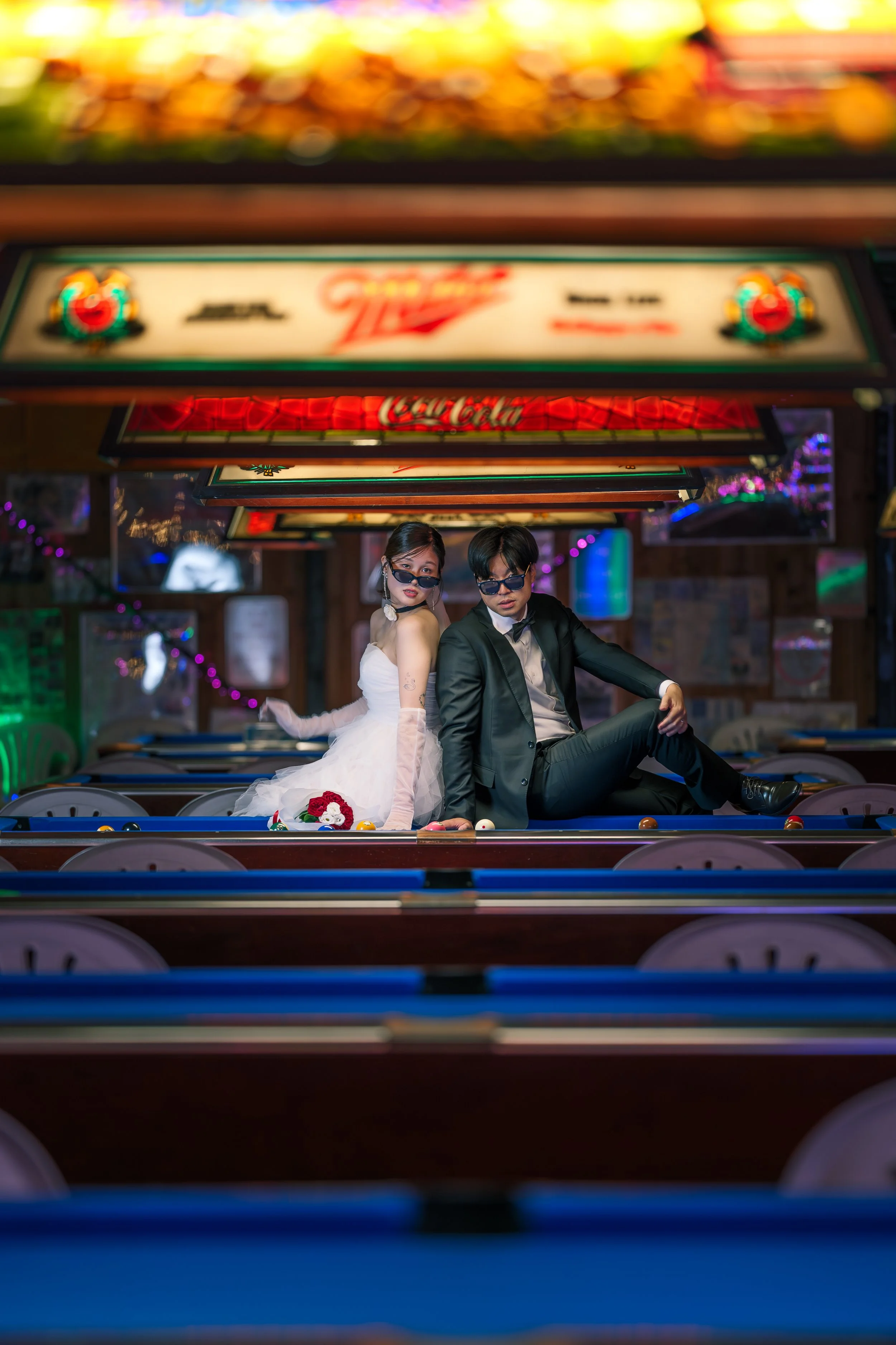 ビリヤード台の上でスーツを着た男性とウェディングドレスの女性がポーズをとっている。背景にはネオンサインやカラフルなライトがある屋内の遊戯場のような場所。